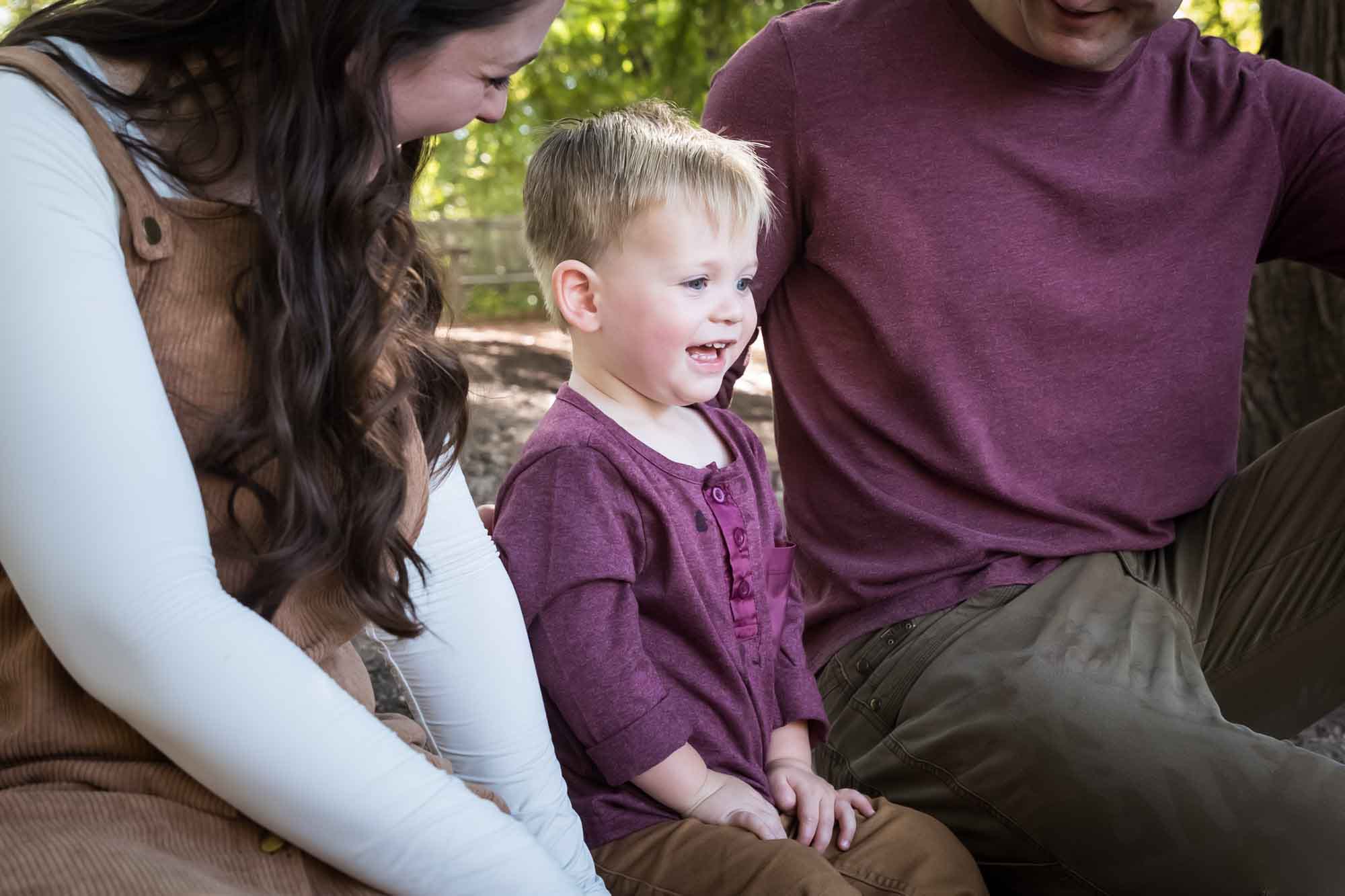 San Antonio Botanical Gardens family portrait of little boy smiling while wearing maroon shirt sitting beside man and woman in the forest