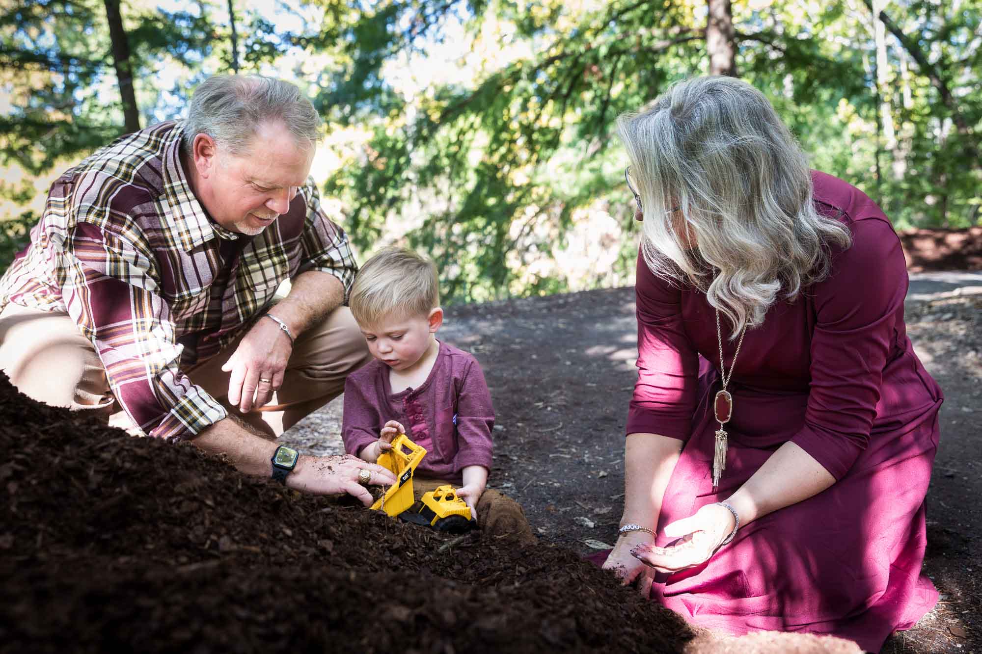 San Antonio Botanical Gardens family portrait of little boy wearing maroon shirt and khaki pants playing with mulch in his toy dump truck alongside his grandparents