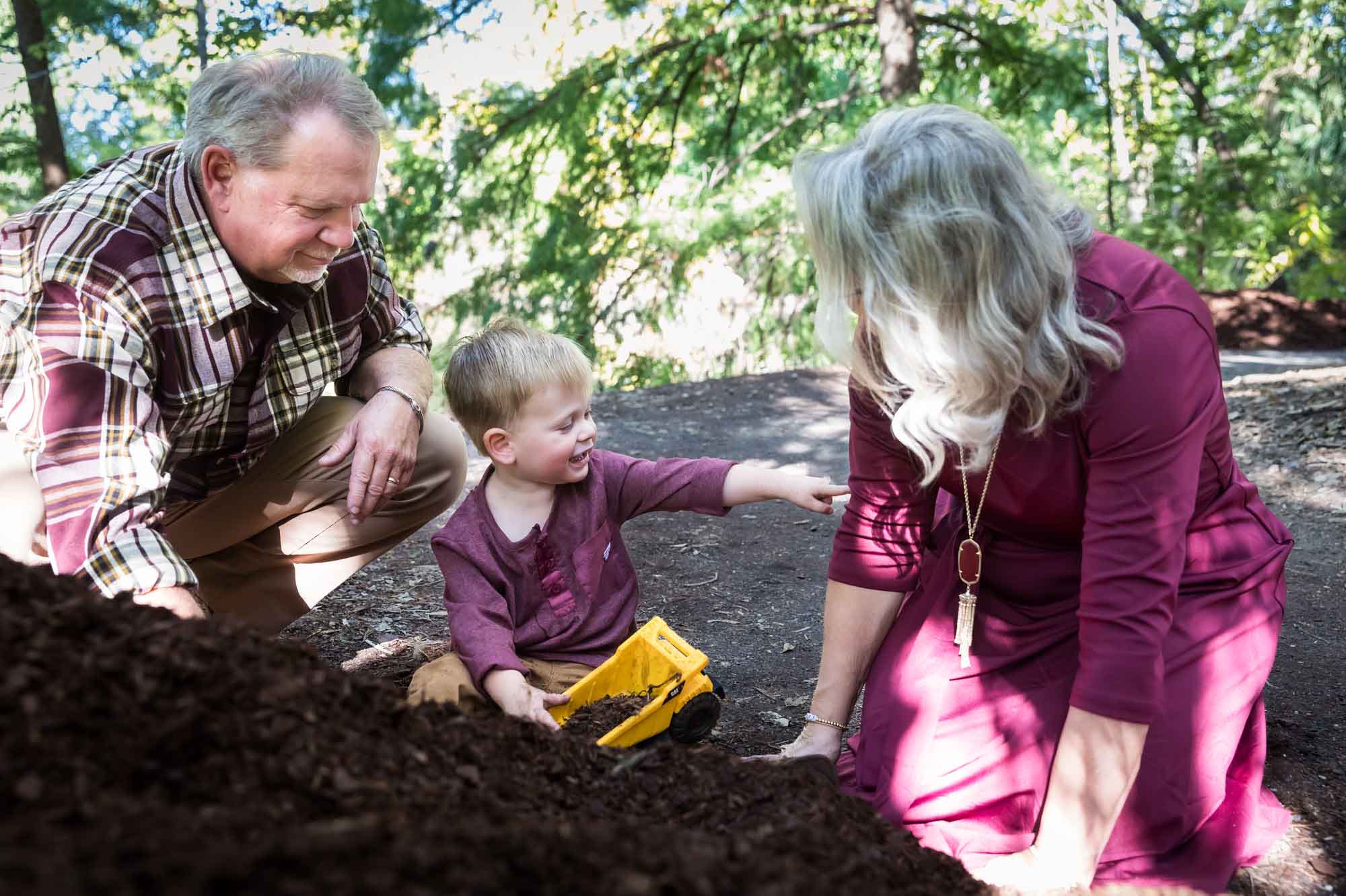San Antonio Botanical Gardens family portrait of little boy wearing maroon shirt and khaki pants playing with mulch in his toy dump truck alongside his grandparents