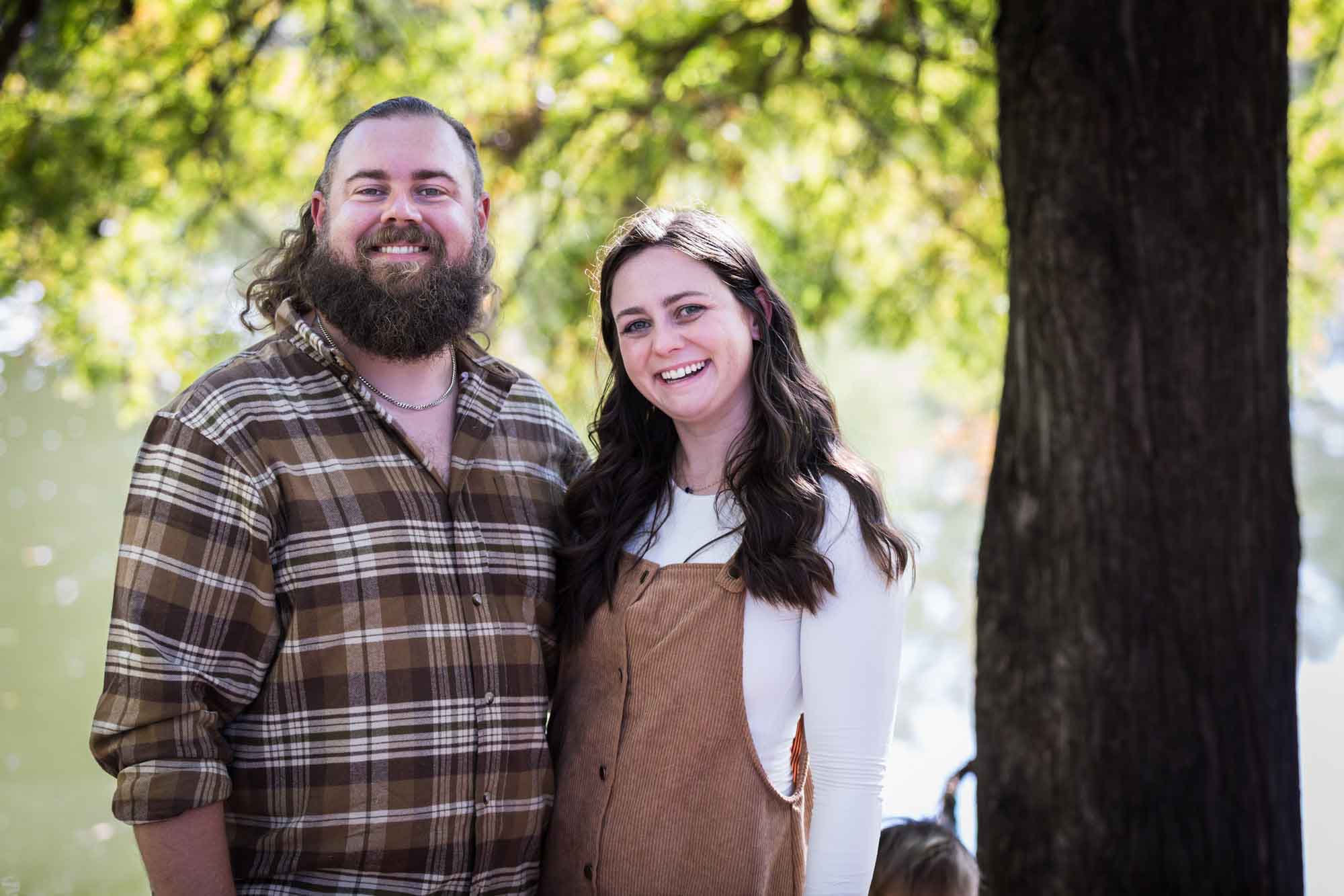 Brother with beard wearing brown plaid shirt standing with sister wearing white shirt and corduroy dress in front of trees during a San Antonio Botanical Gardens family portrait session