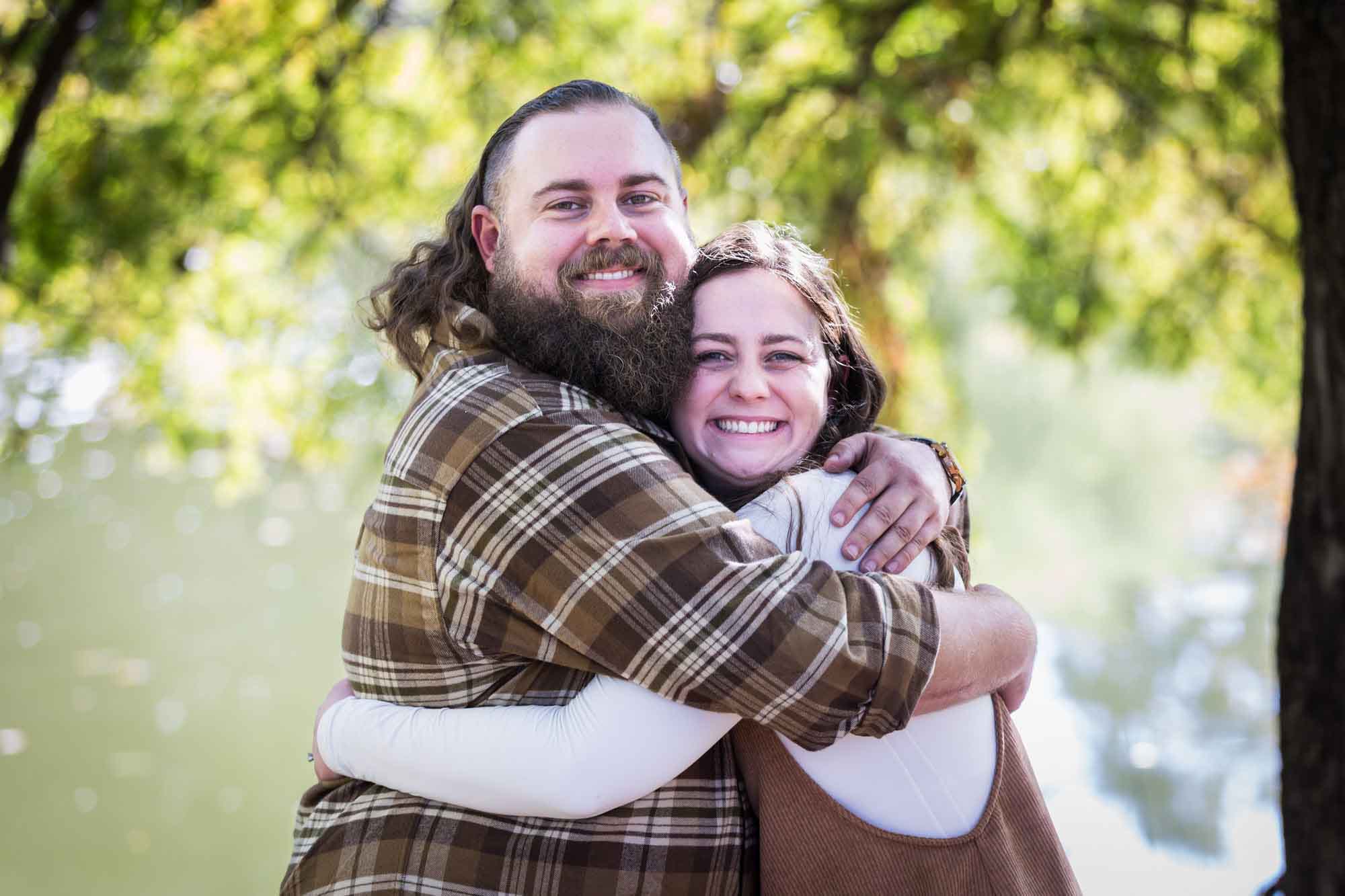 Brother with beard wearing brown plaid shirt hugging sister wearing white shirt and corduroy dress in front of trees during a San Antonio Botanical Gardens family portrait session
