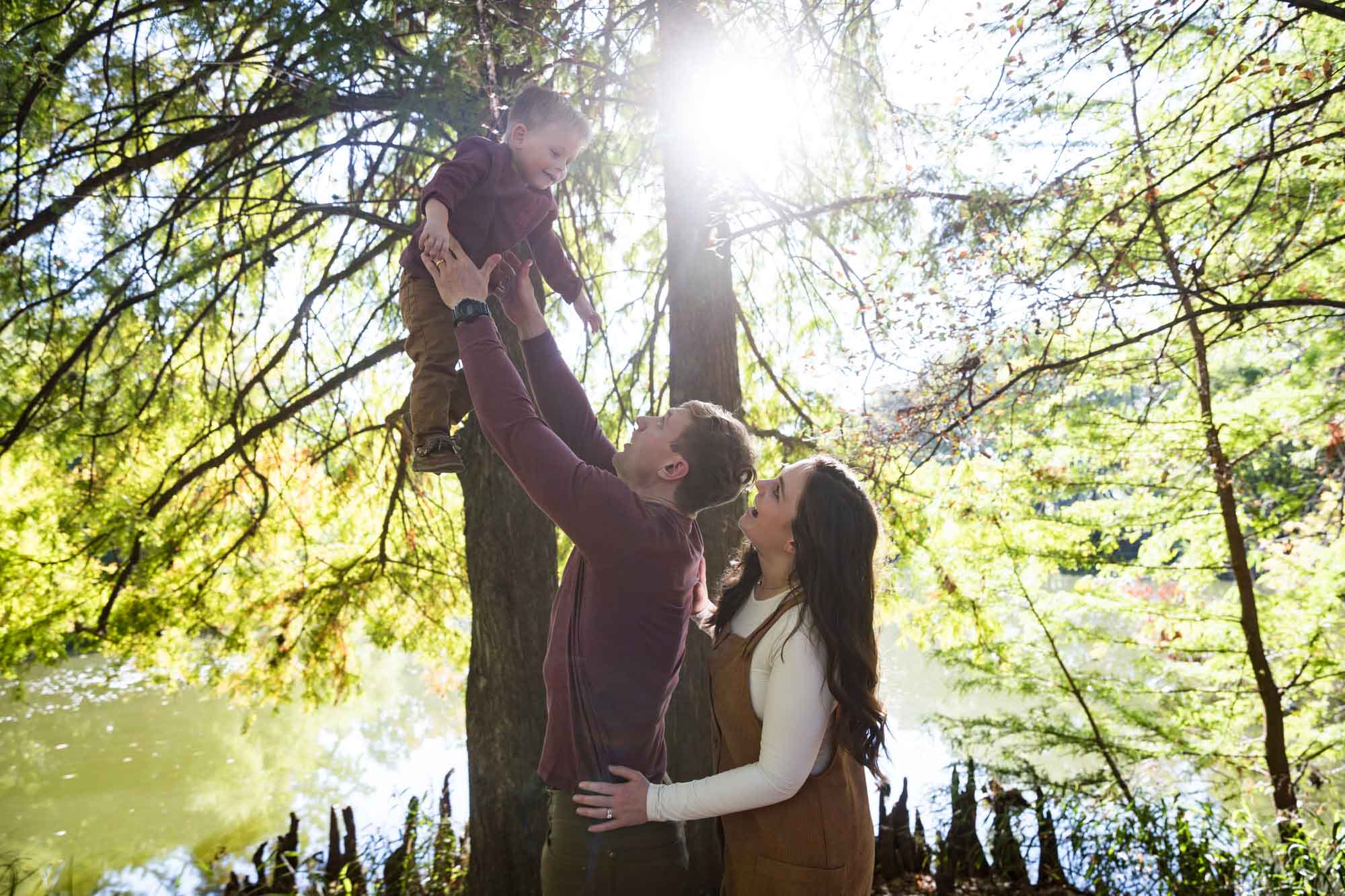 Mother and father looking at little boy being held up in the air in front of trees in front of lake during a San Antonio Botanical Gardens family portrait session