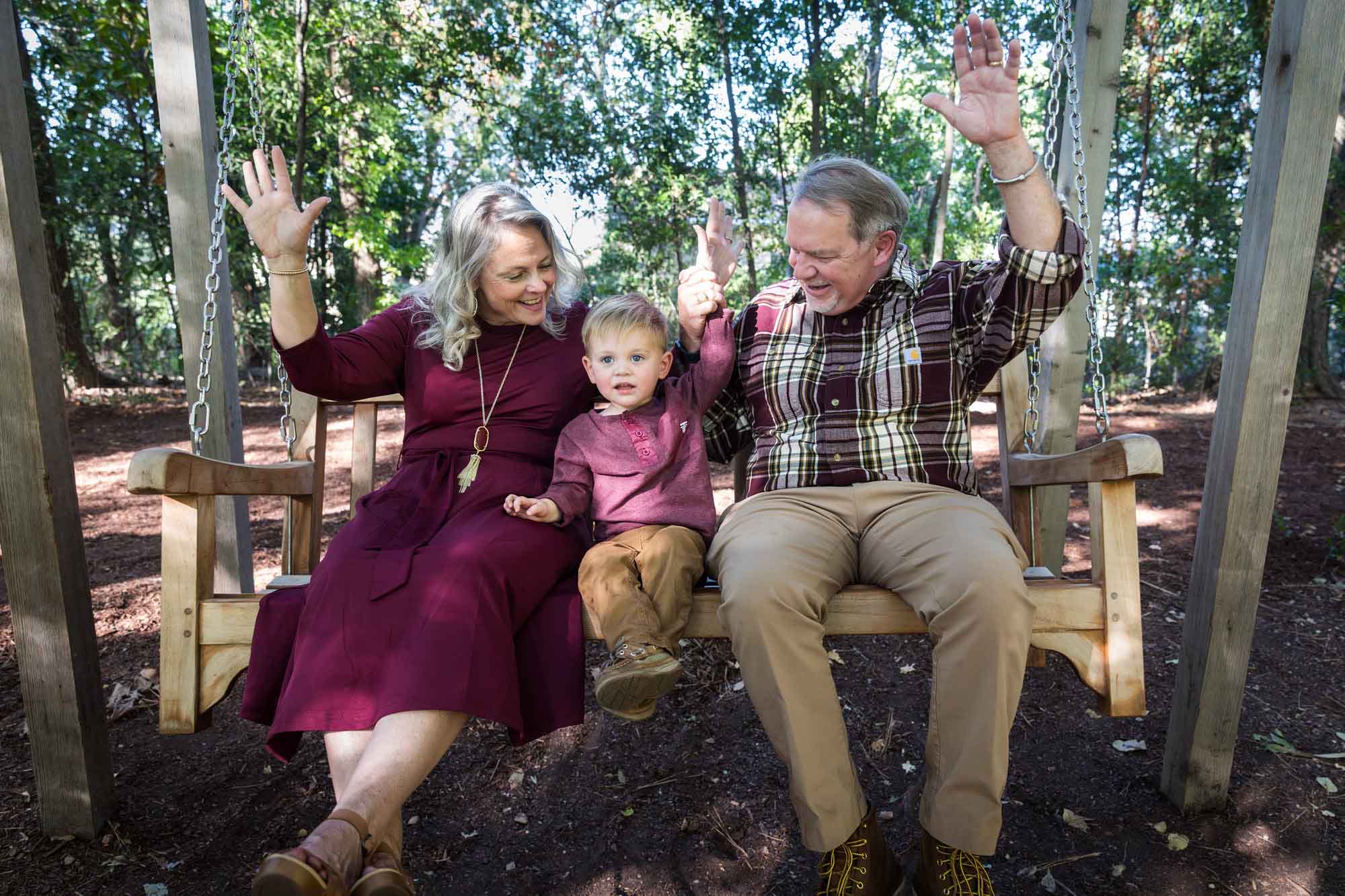 Grandparents with hands up in the air swinging on bench swing with little boy in forest during a San Antonio Botanical Gardens family portrait session