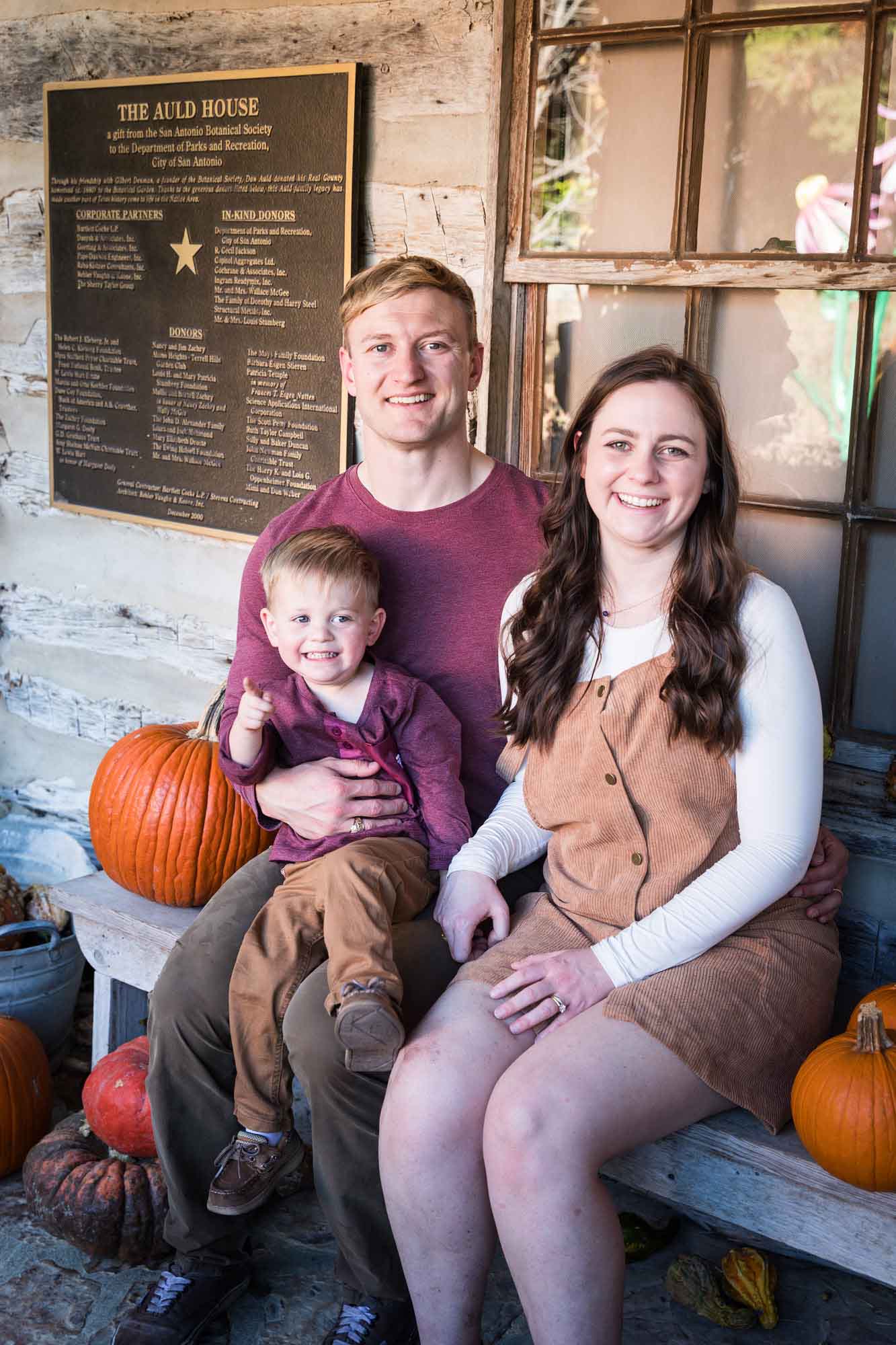 Mother and father holding little boy holding small pumpkin seated on bench in front of log cabin wall during a San Antonio Botanical Gardens family portrait session