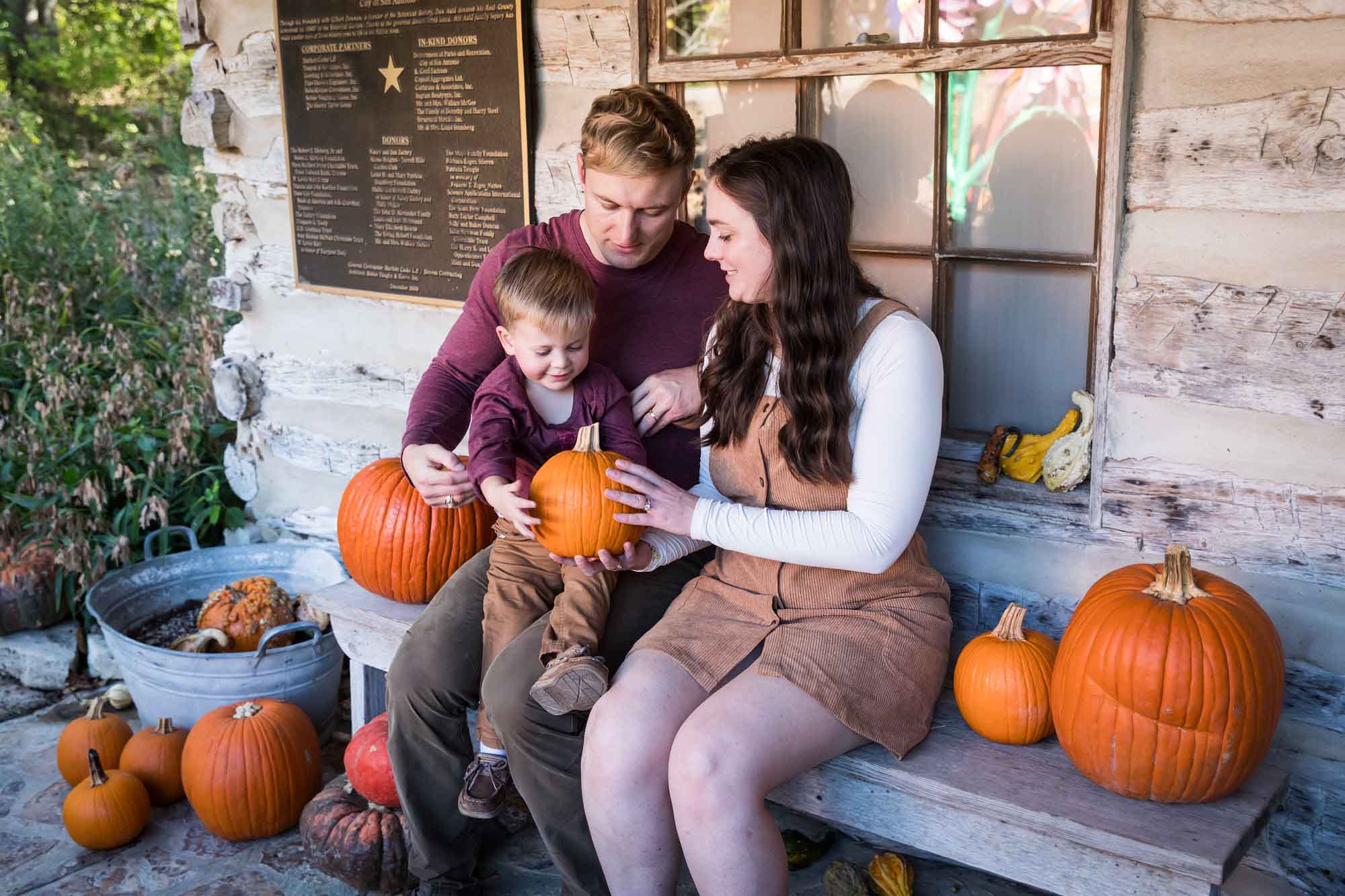 Mother and father holding little boy holding small pumpkin seated on bench in front of log cabin wall during a San Antonio Botanical Gardens family portrait session