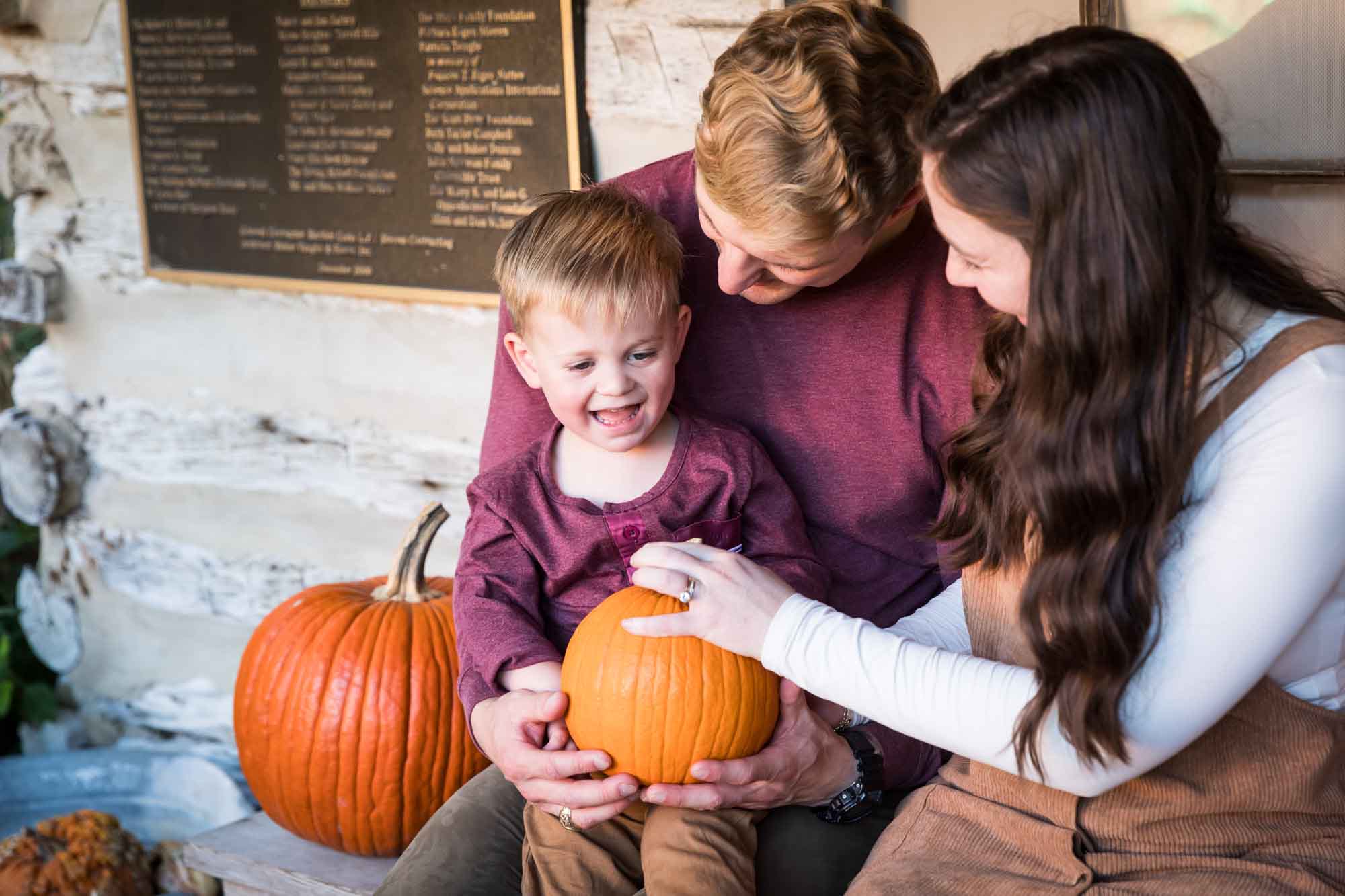 Mother and father holding little boy holding small pumpkin in front of log cabin wall during a San Antonio Botanical Gardens family portrait session