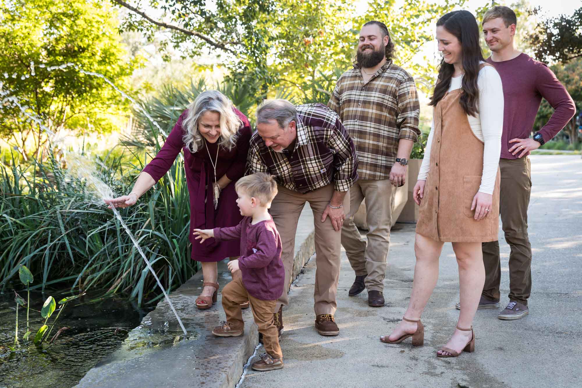 Little boy playing in water stream with grandparents and family in front of bushes during a San Antonio Botanical Gardens family portrait session