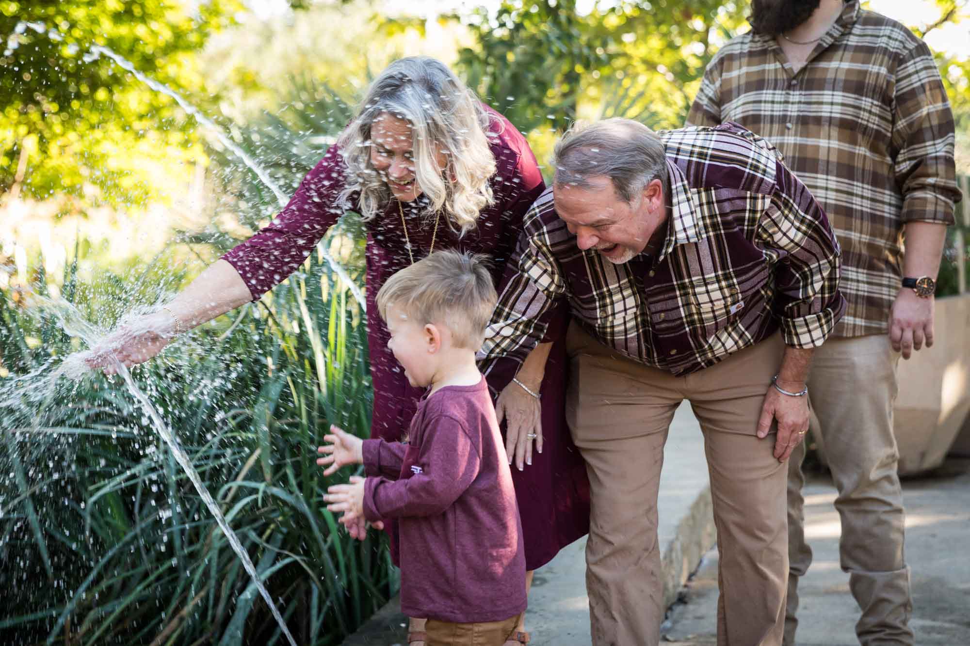 Little boy playing in water stream with grandparents and family in front of bushes during a San Antonio Botanical Gardens family portrait session
