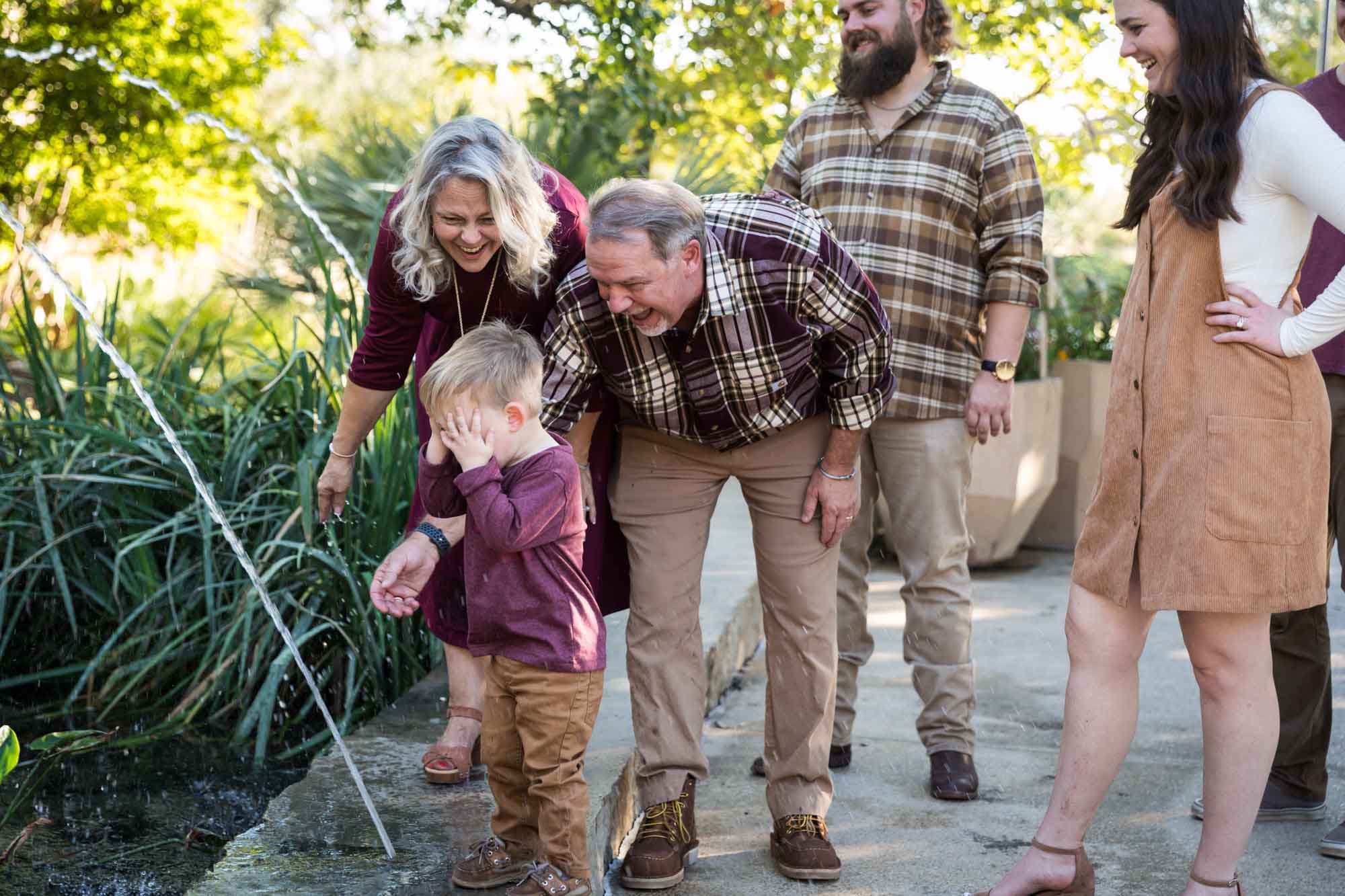 Little boy playing in water stream with grandparents and family in front of bushes during a San Antonio Botanical Gardens family portrait session
