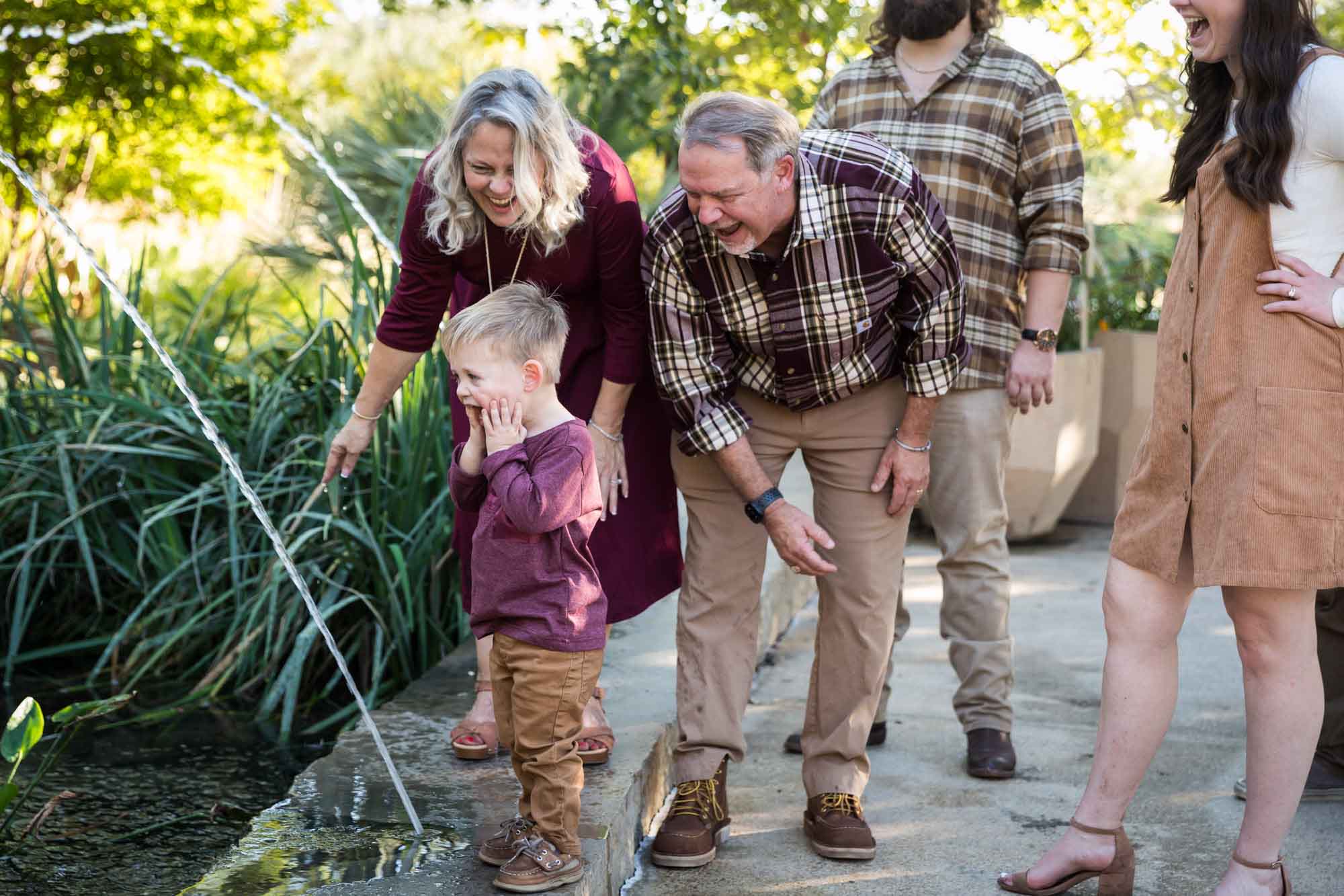 Little boy playing in water stream with grandparents and family in front of bushes during a San Antonio Botanical Gardens family portrait session