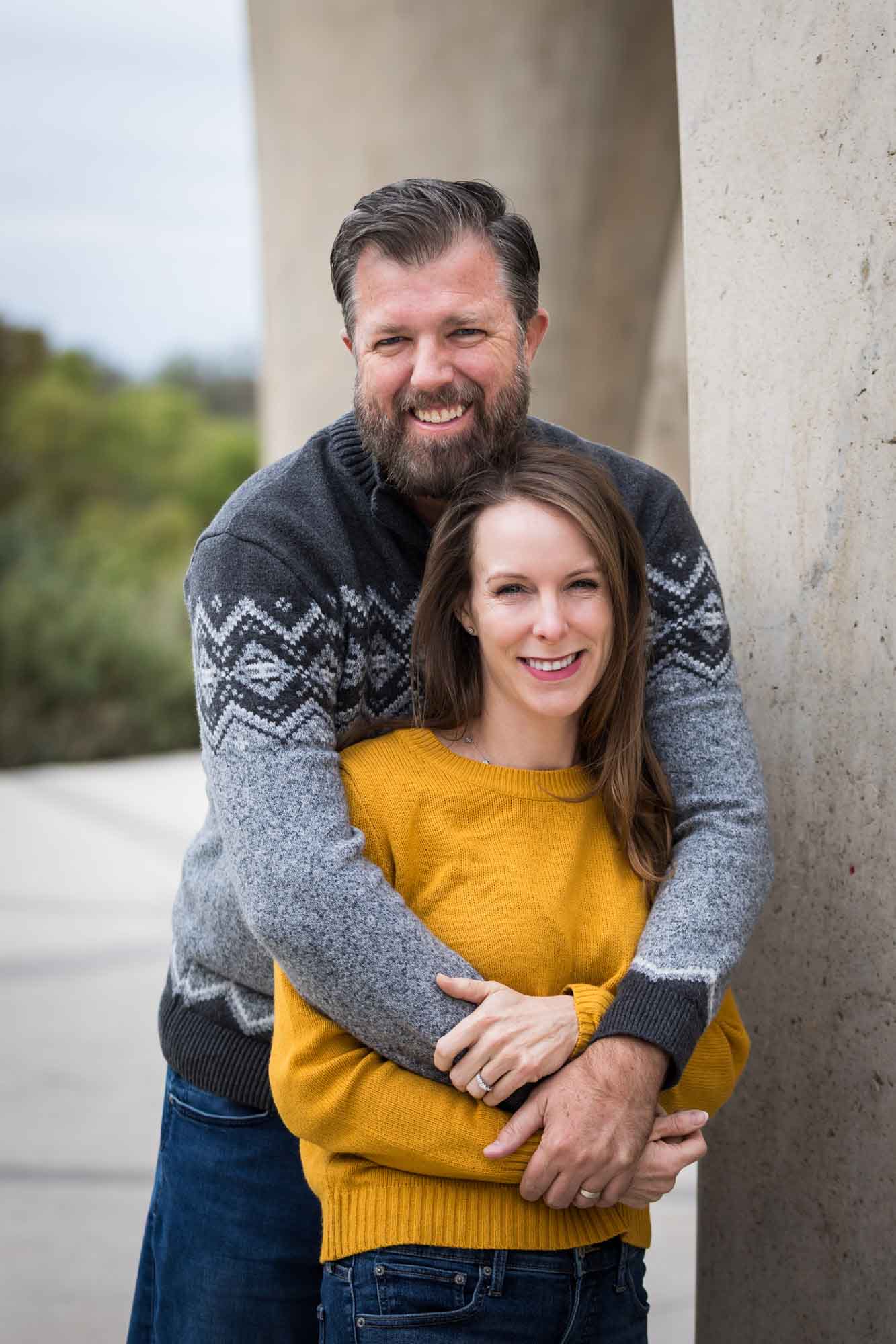 Man wearing grey sweater hugging woman wearing yellow sweater and jeans during a Confluence Park family portrait session