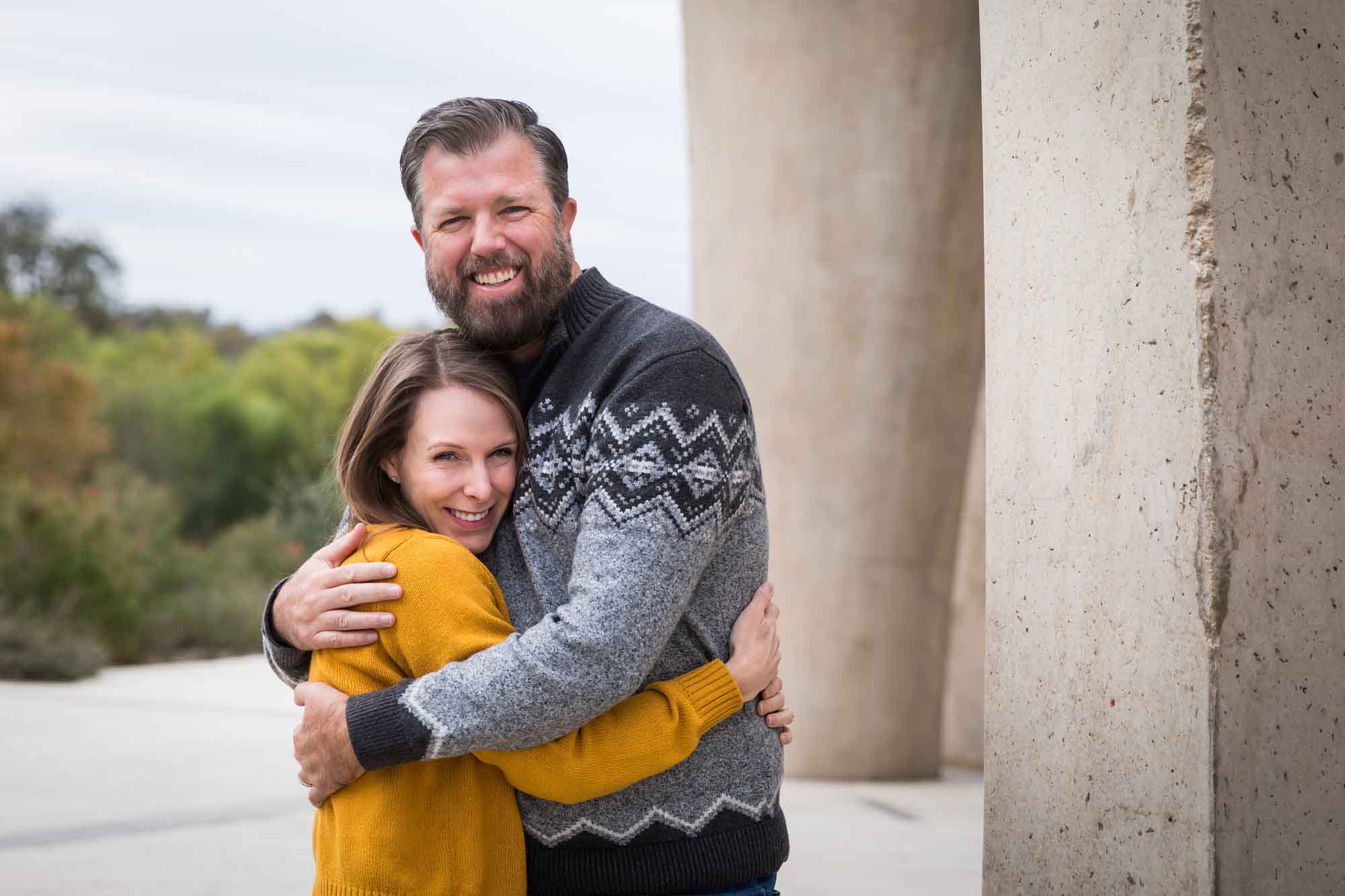 Man wearing grey sweater hugging woman wearing yellow sweater and jeans during a Confluence Park family portrait session
