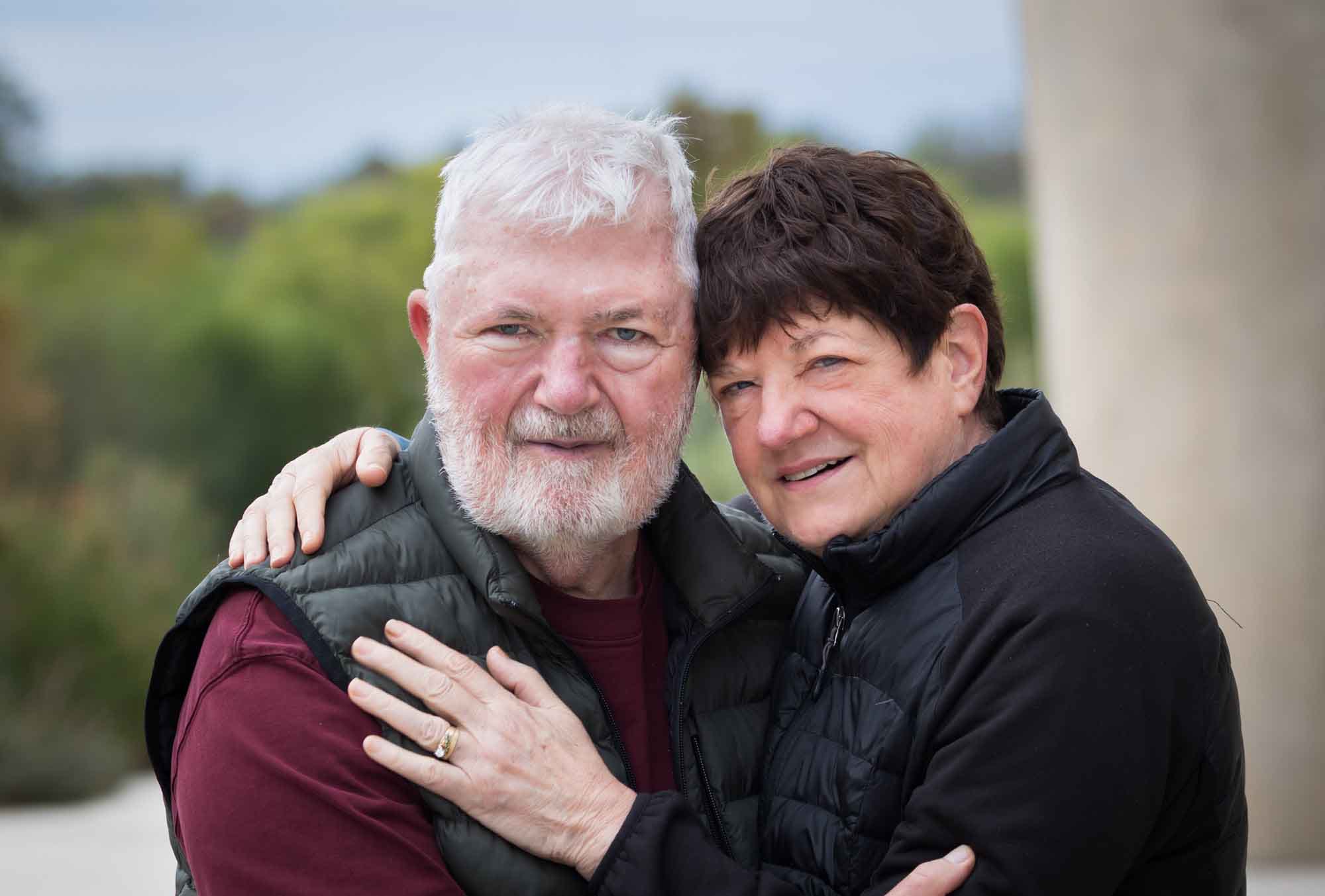 Older man wearing puffy vest and maroon shirt hugging an older woman wearing black sweater during a Confluence Park family portrait session