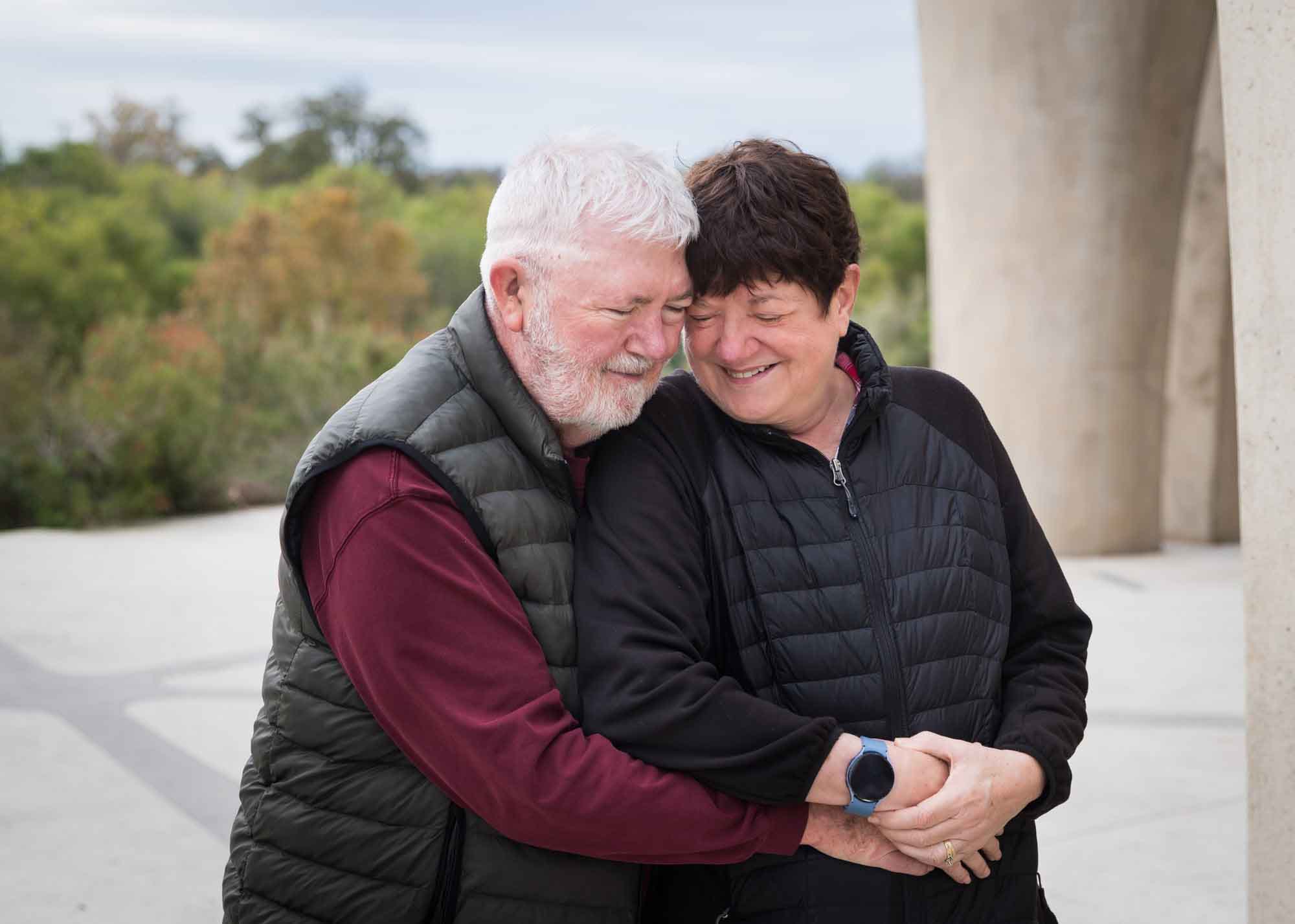 Older man wearing puffy vest and maroon shirt hugging an older woman wearing black sweater during a Confluence Park family portrait session