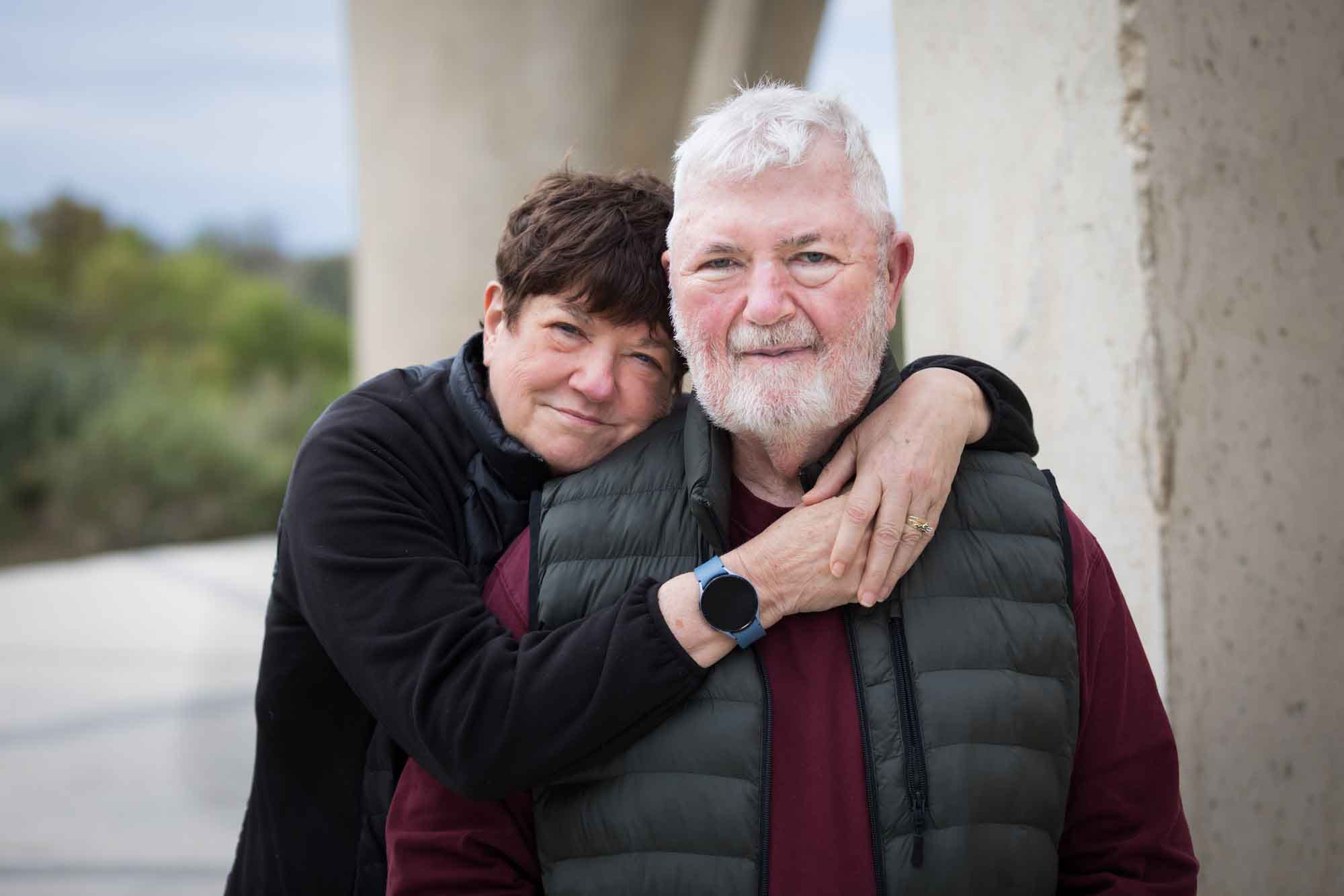 Older man wearing puffy vest and maroon shirt being hugged by older woman wearing black sweater during a Confluence Park family portrait session