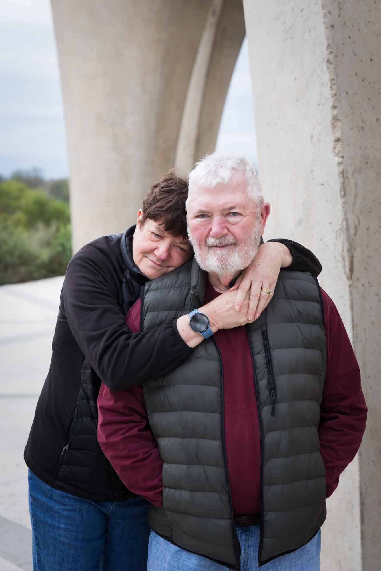 Older man wearing puffy vest and maroon shirt being hugged by older woman wearing black sweater during a Confluence Park family portrait session