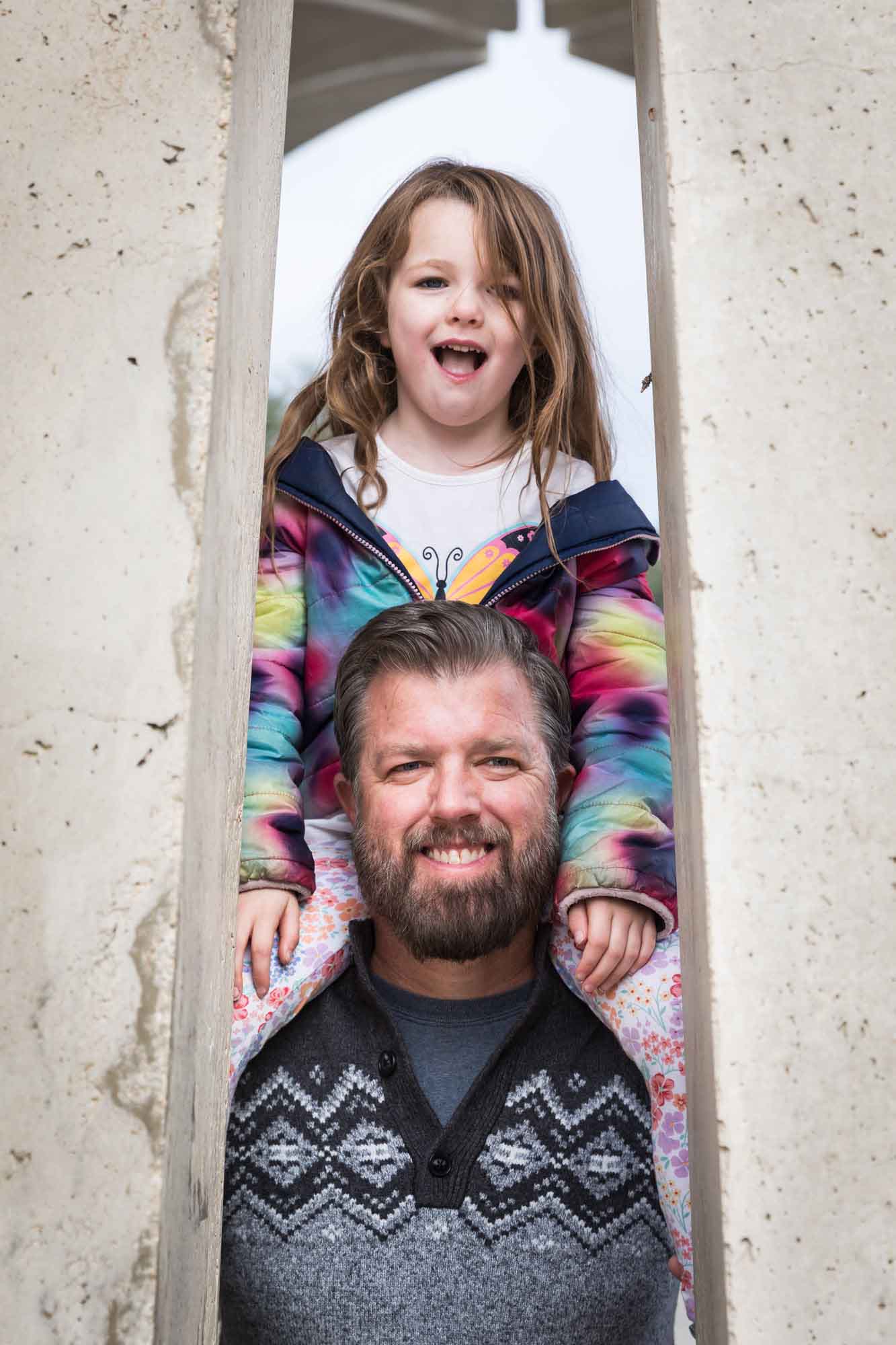 Father wearing grey sweater holding little girl wearing multicolored sweater on his shoulders during a Confluence Park family portrait session