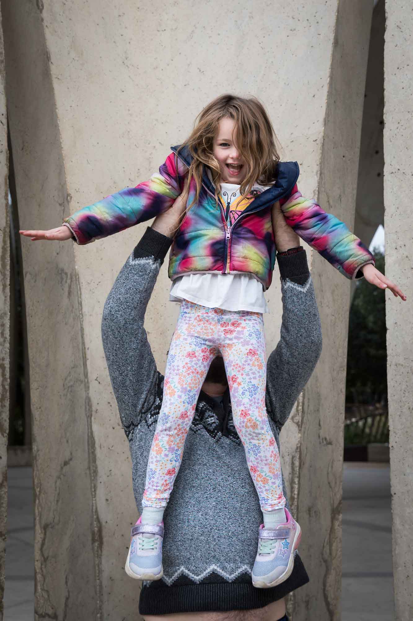Father wearing grey sweater holding up a little girl wearing multicolored sweater during a Confluence Park family portrait session