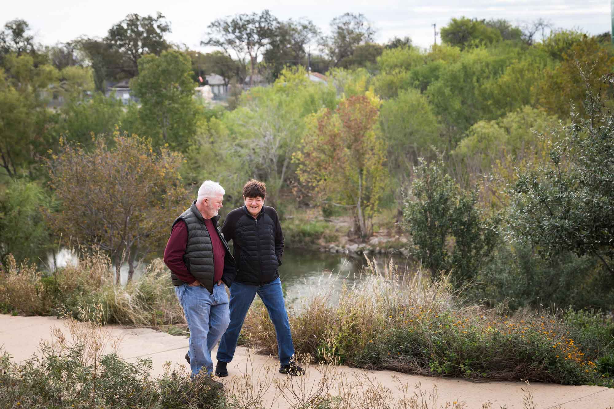 Older man wearing puffy vest and maroon shirt walking with an older woman wearing black sweater down a park pathway during a Confluence Park family portrait session