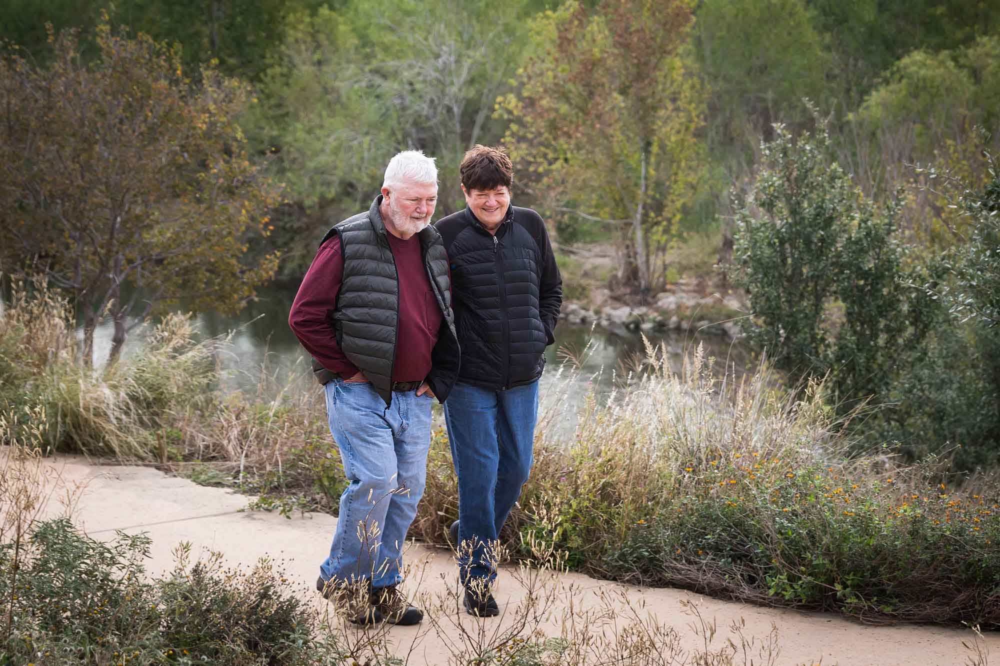 Older man wearing puffy vest and maroon shirt walking with an older woman wearing black sweater down a park pathway during a Confluence Park family portrait session