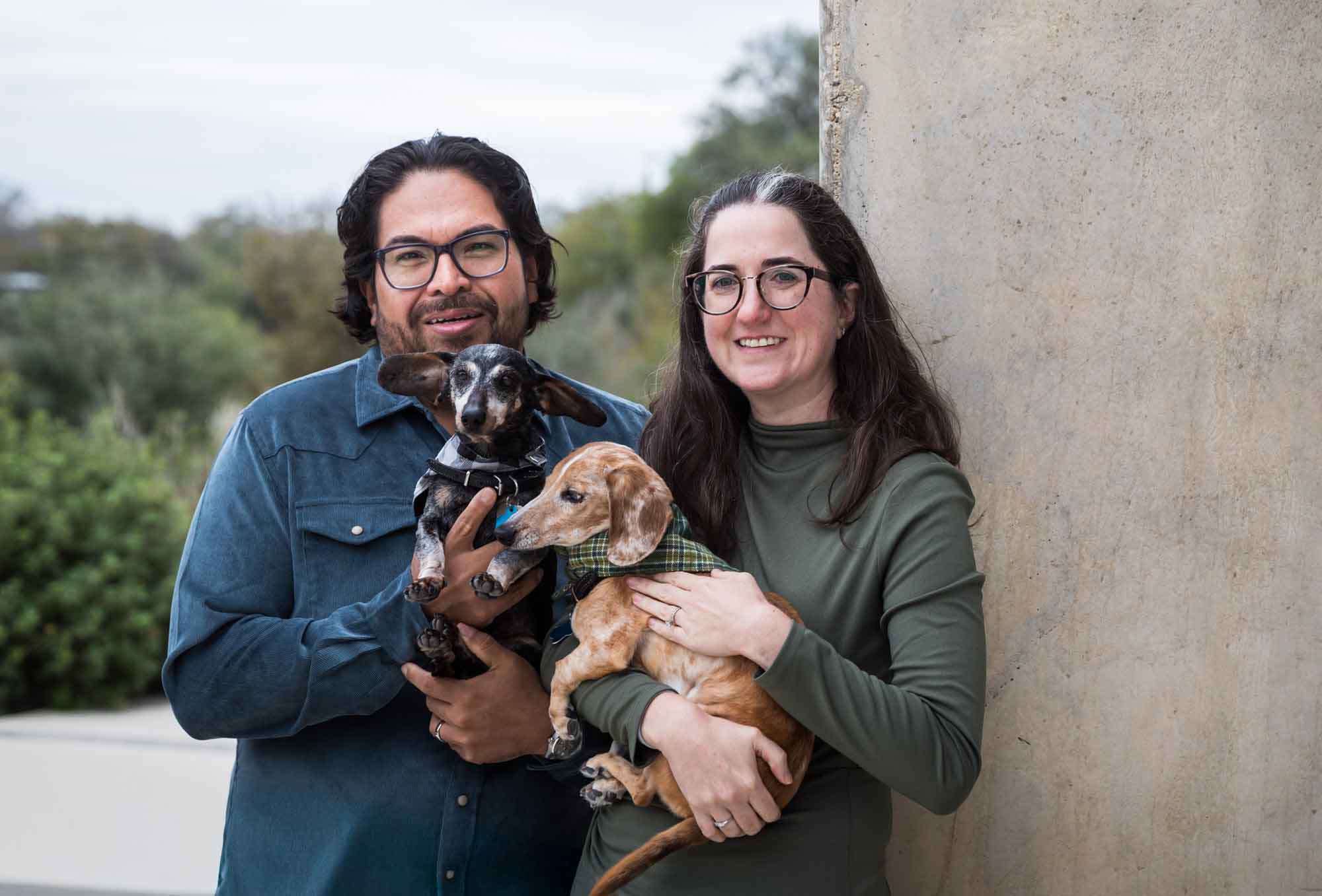 Man wearing denim long-sleeved shirt standing beside woman wearing green long-sleeved shirt holding two dachshunds during a Confluence Park family portrait session