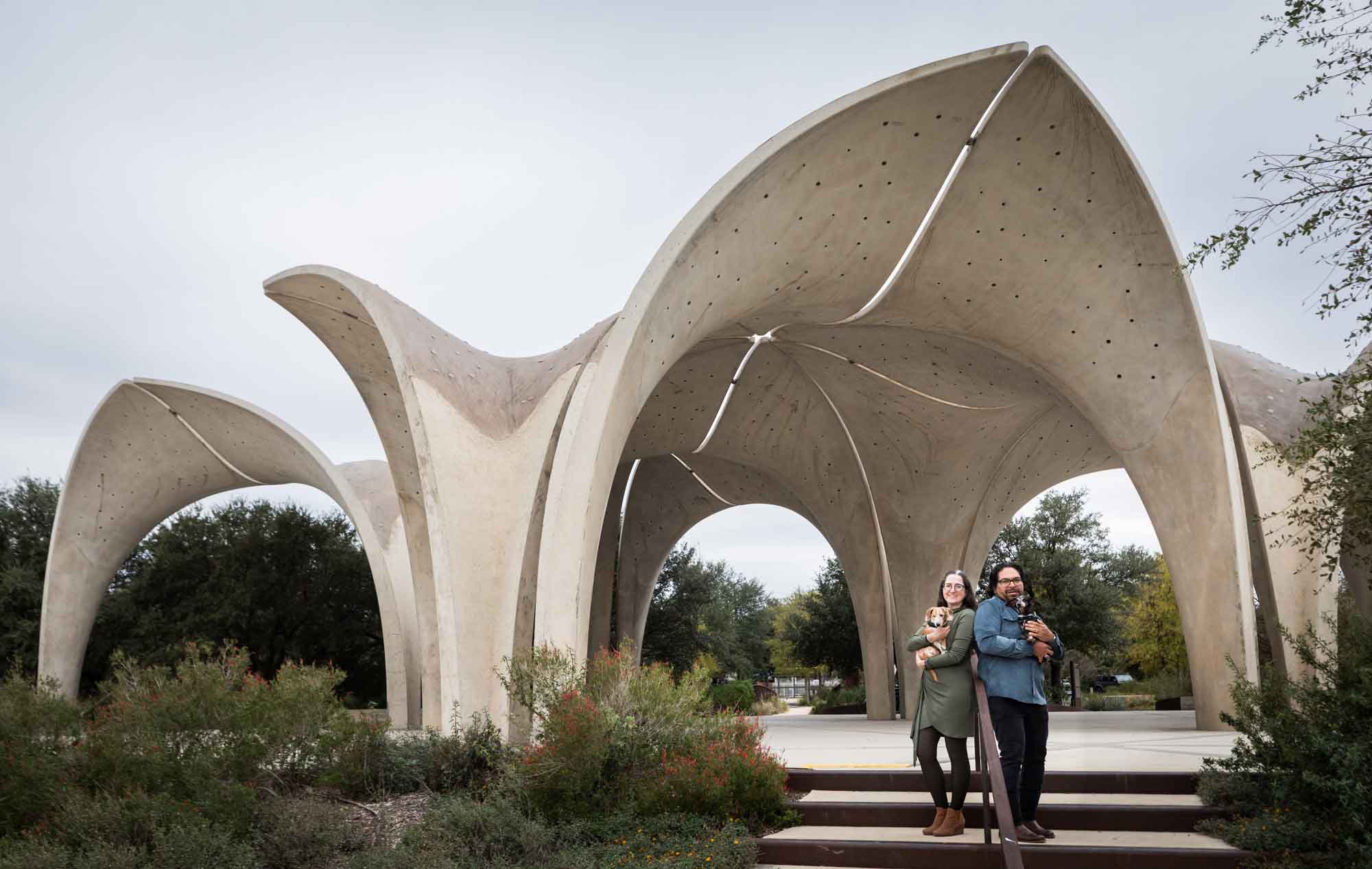 Man wearing denim long-sleeved shirt and a woman wearing green long-sleeved shirt standing together holding two dachshunds on stairs under concrete arch during a Confluence Park family portrait session