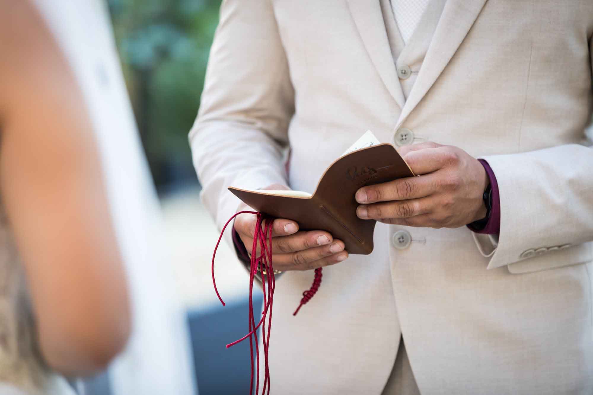 Close up of groom holding brown vow book and red fabric bracelet during ceremony at a San Antonio DIY wedding