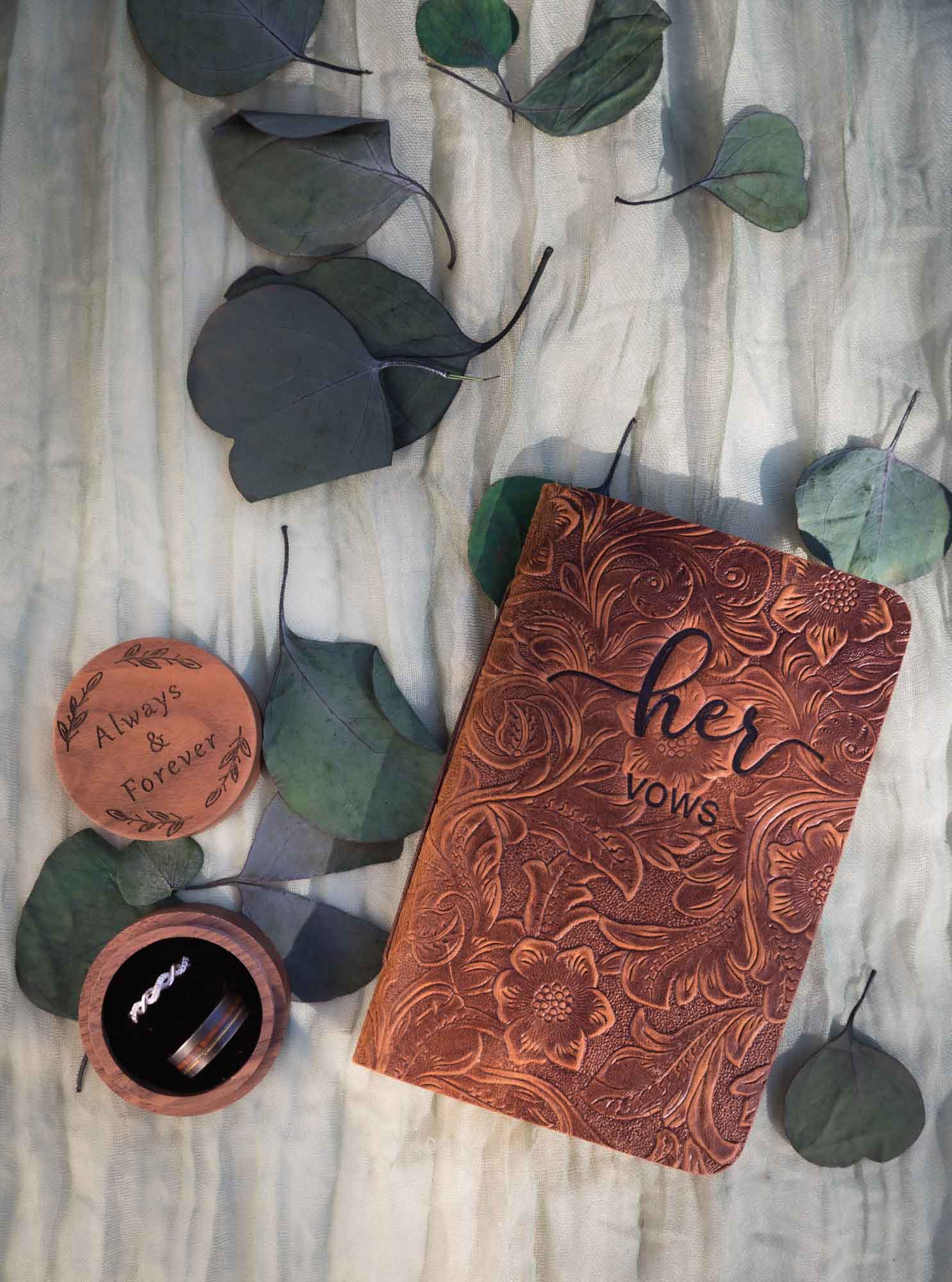 Leather vow book, round ring box on table with eucalyptus leaves at a San Antonio DIY wedding