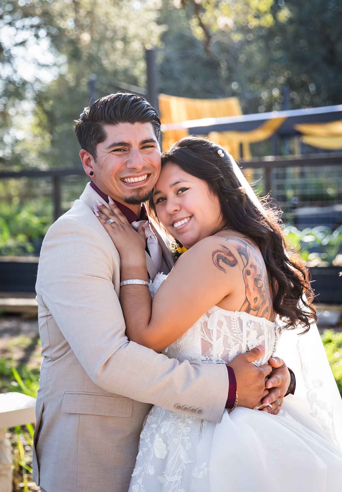 Bride in strapless gown and veil hugging groom wearing tan suit in front of large oak tree at a San Antonio DIY wedding