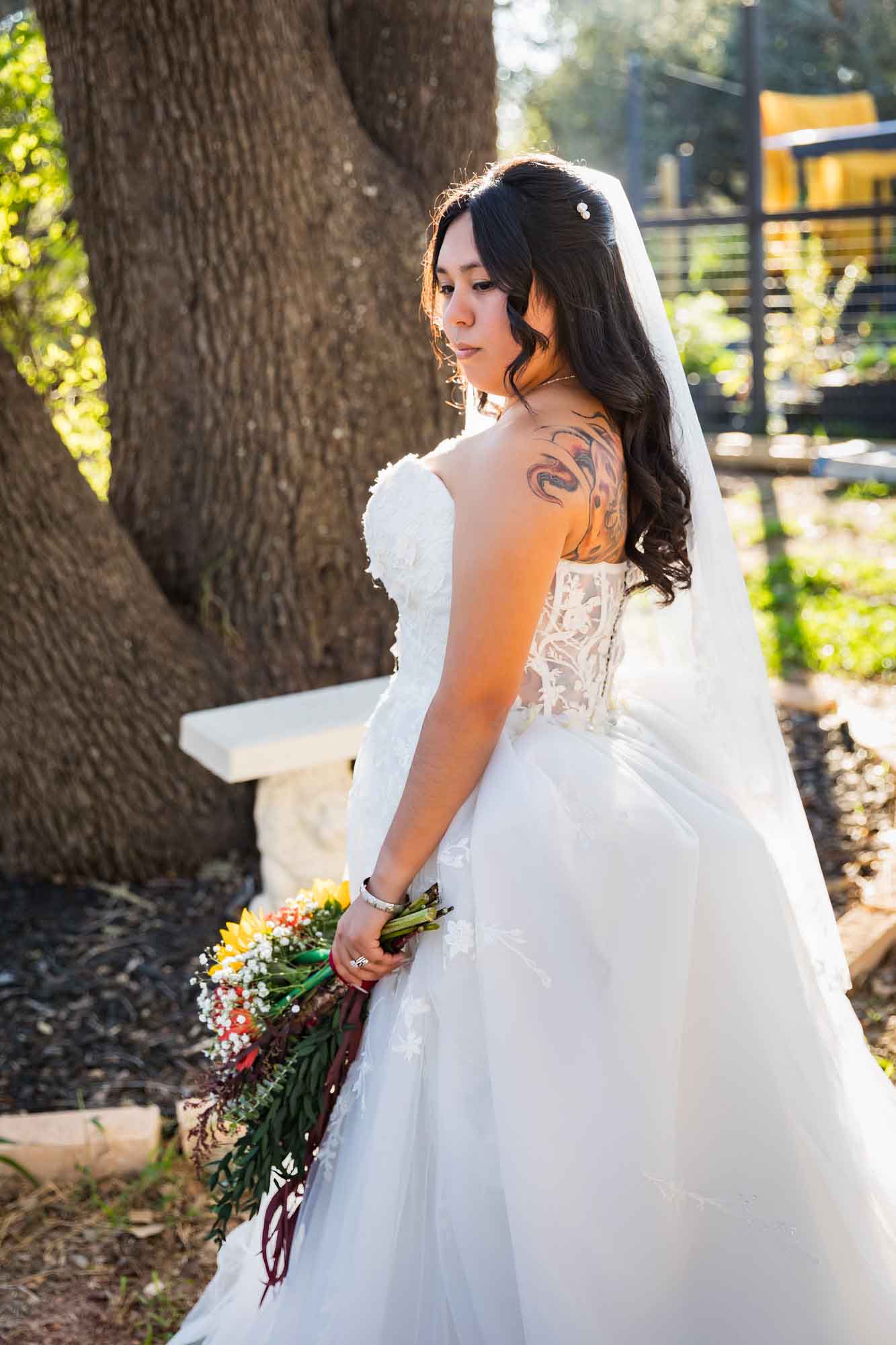Bride wearing strapless white gown and veil with her back to the camera holding flowers standing in front of oak tree at a San Antonio DIY wedding