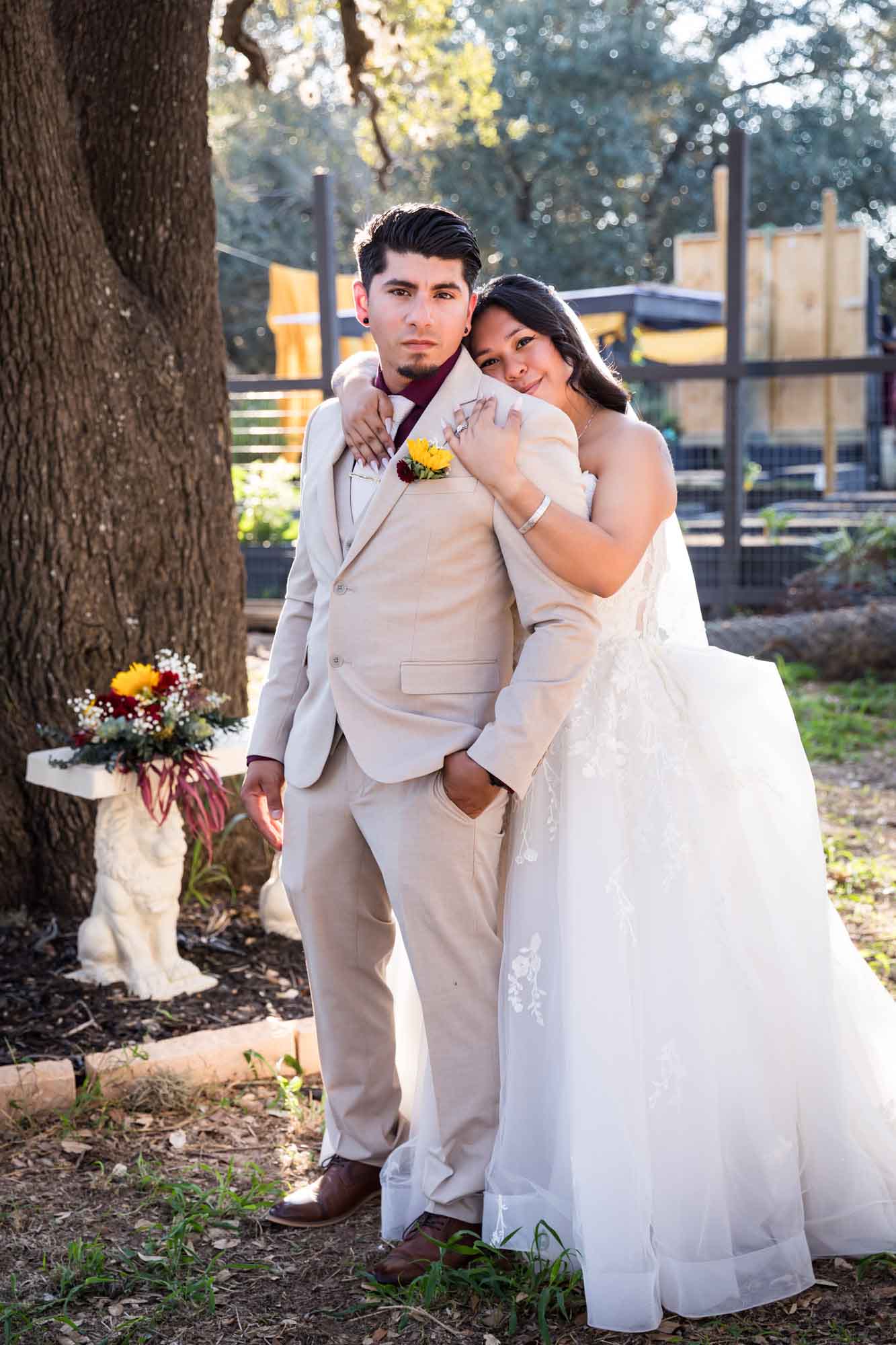 Bride in strapless gown and veil hugging groom wearing tan suit in front of large oak tree at a San Antonio DIY wedding