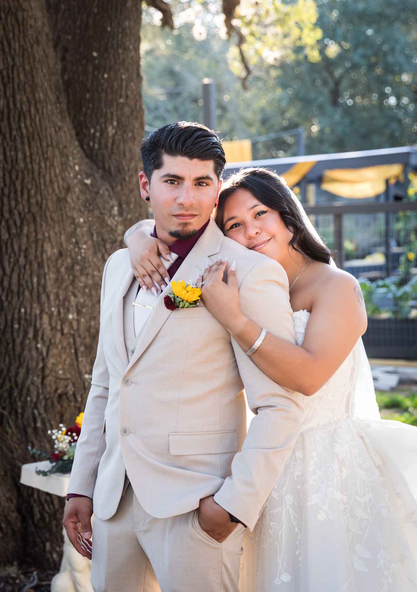 Bride in strapless gown and veil hugging groom wearing tan suit in front of large oak tree at a San Antonio DIY wedding