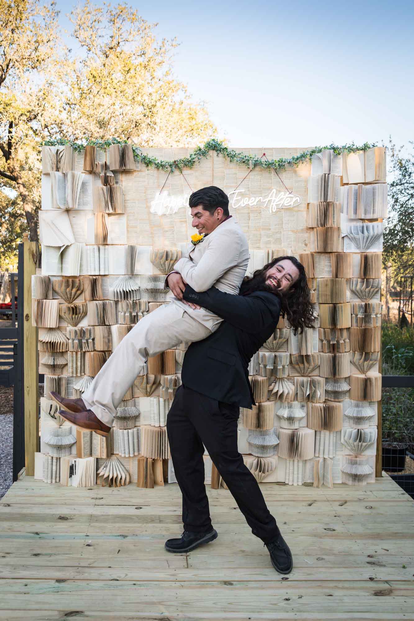 Man in black suit lifting up groom in tan suit in front of book background at a San Antonio DIY wedding