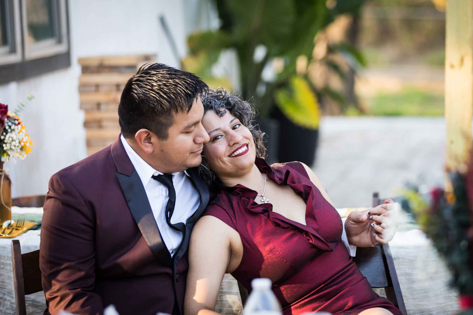 Man wearing maroon suit sitting closely with woman searing sleeveless dark red dress at reception at a San Antonio DIY wedding