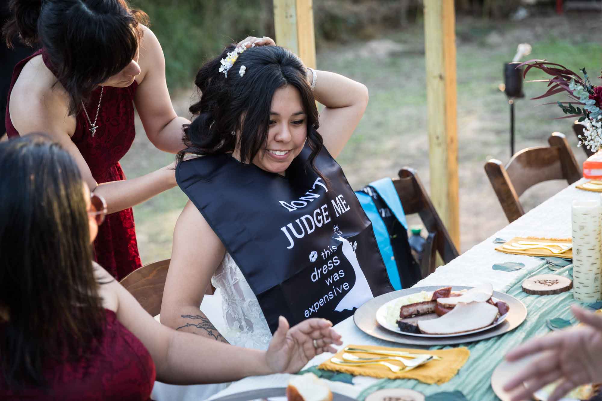 Bride seated during reception while female guest puts black bib on bride at a San Antonio DIY wedding