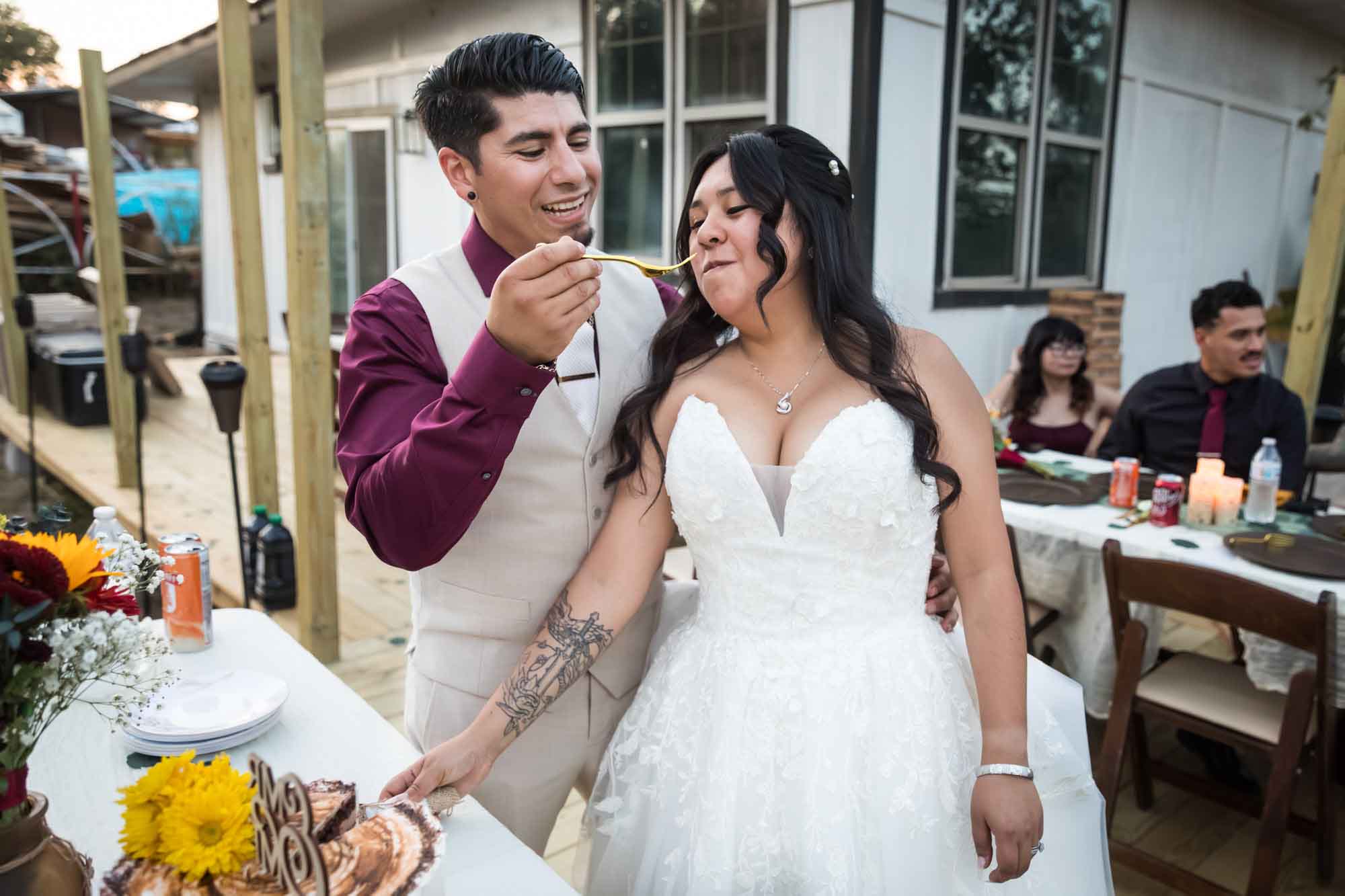 Groom in maroon shirt and tan vest feeding cake to bride in white dress at a San Antonio DIY wedding