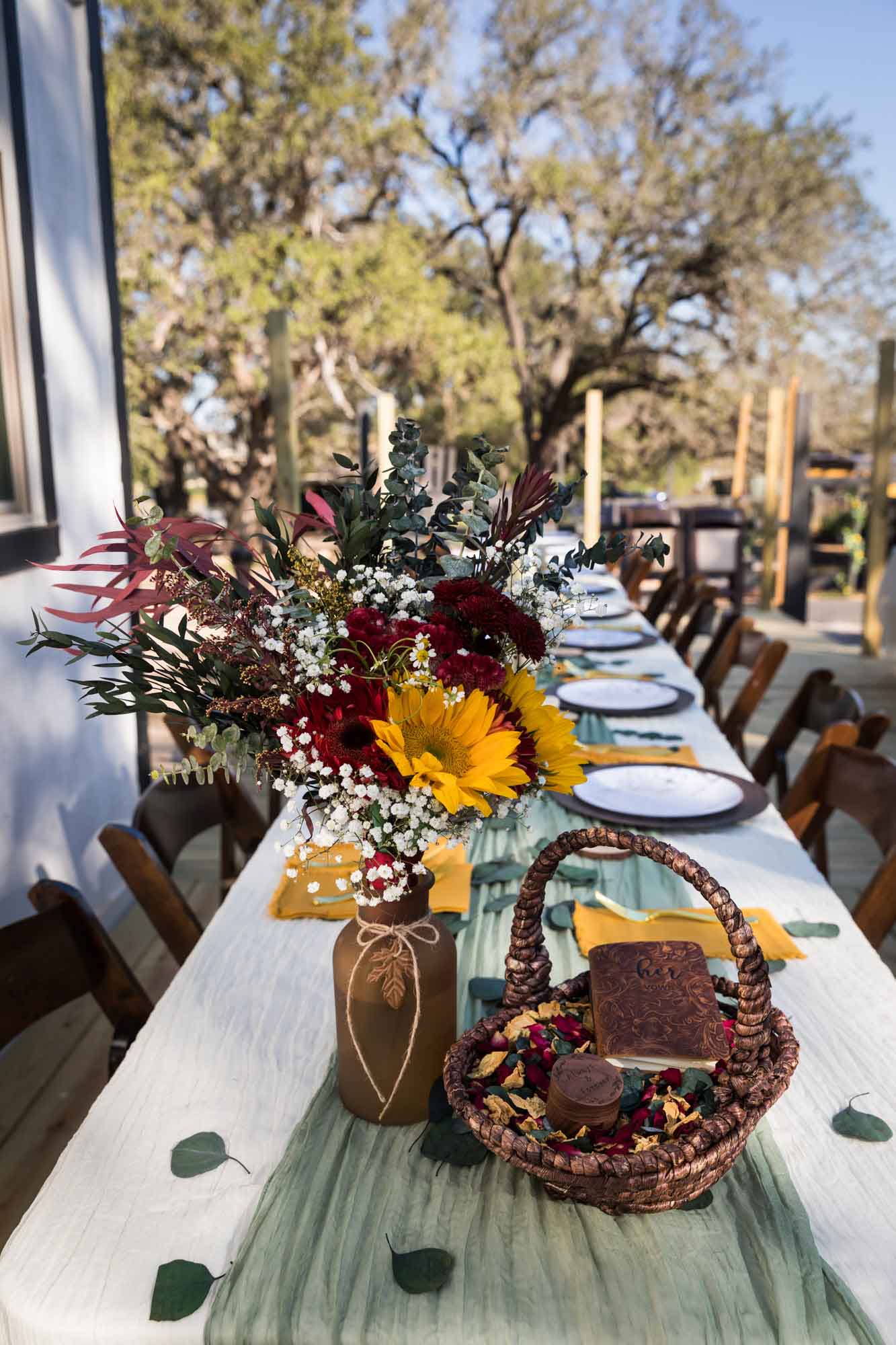 Table with brown glass vase holding multicolored flowers and basket with flower petals and brown vow book at a San Antonio DIY wedding