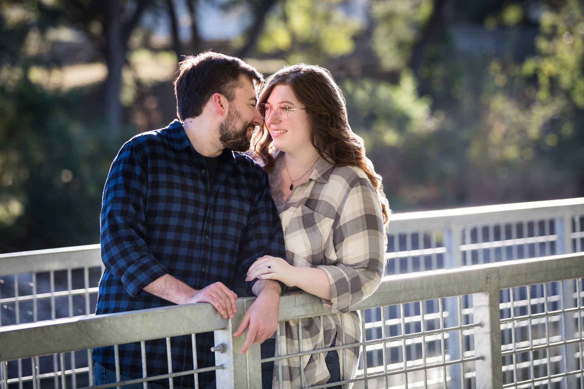 Boerne engagement photos of couple wearing plaid shirts leaning over metal bridge in River Road Park