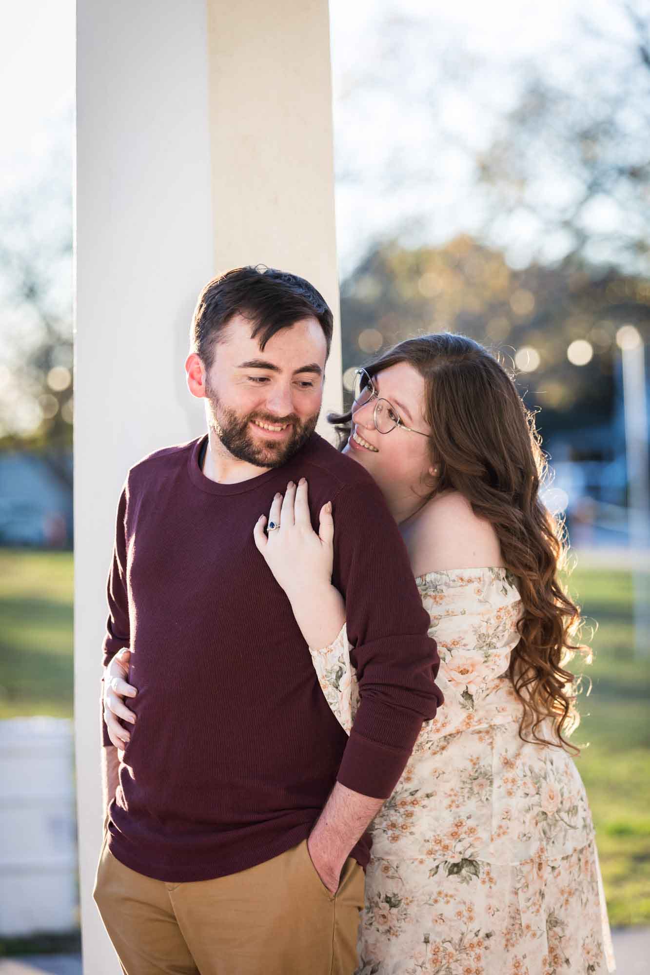 Boerne engagement photos of woman hugging man from behind in front of white column in Main Plaza