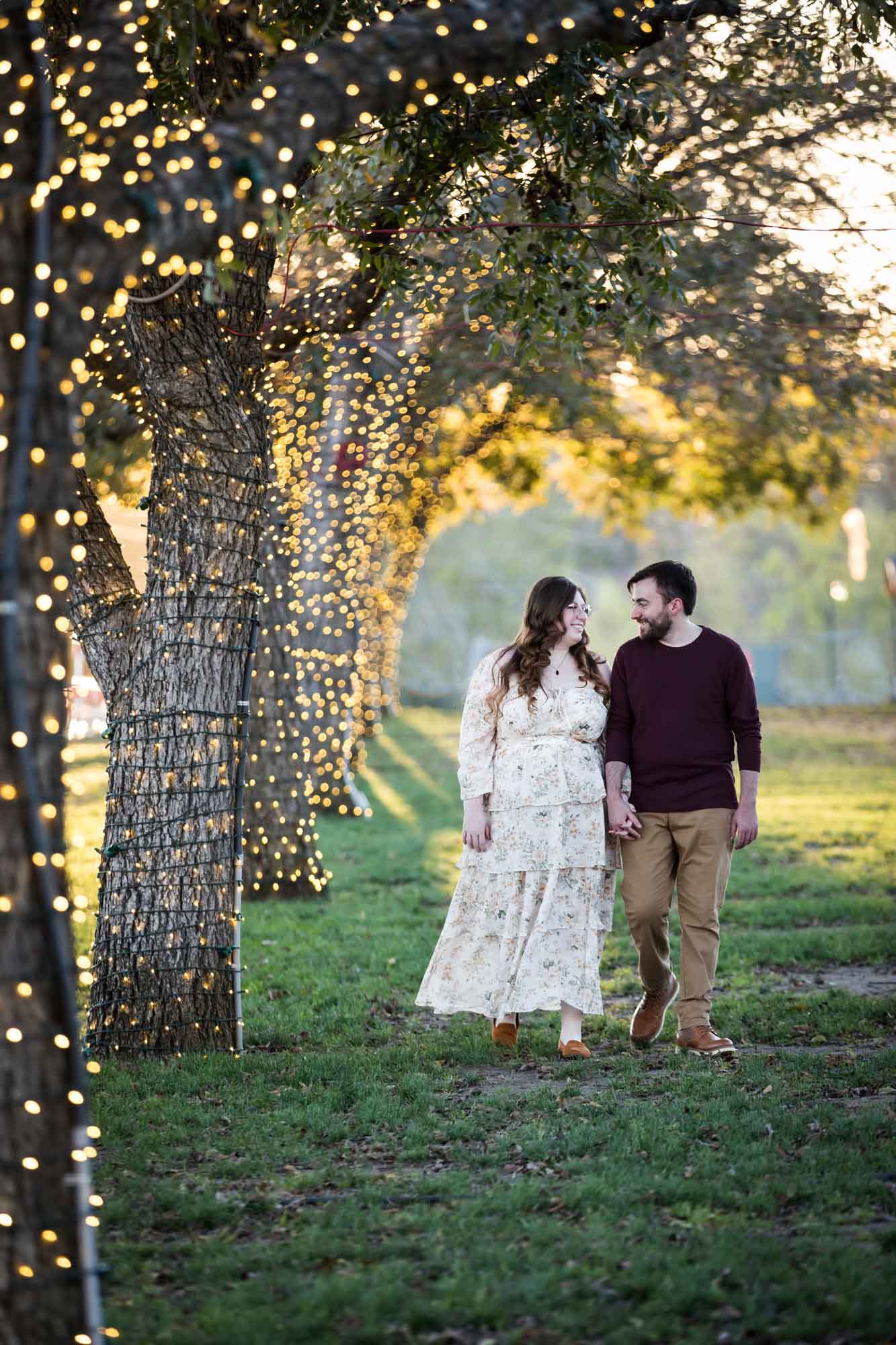 Boerne engagement photos of woman and man walking beside trees wrapped in string lights in Main Plaza