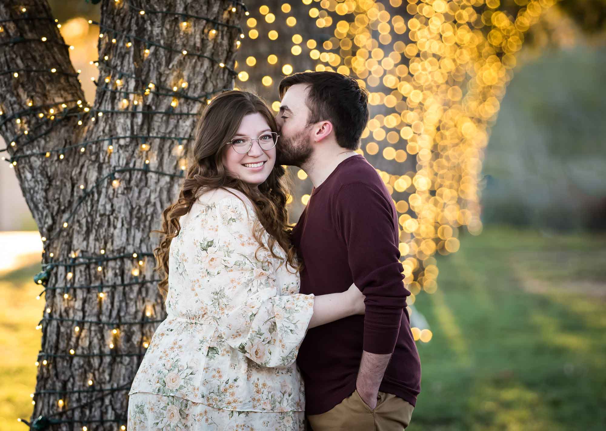Boerne engagement photos of woman and man kissing beside trees wrapped in string lights in Main Plaza