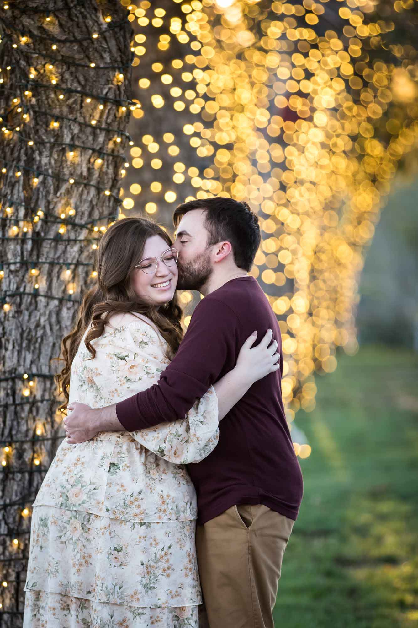 Boerne engagement photos of woman and man kissing beside trees wrapped in string lights in Main Plaza