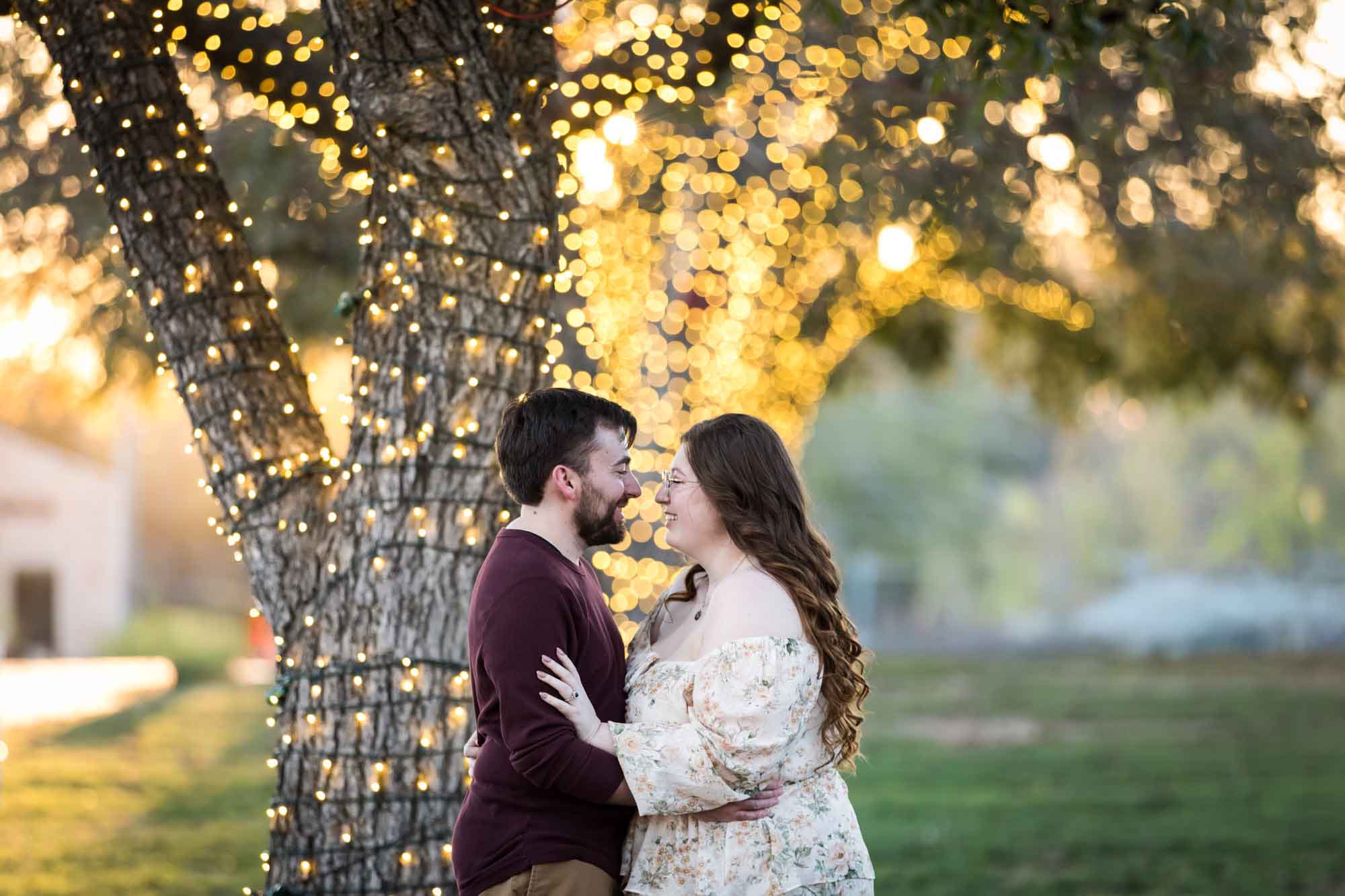 Boerne engagement photos of woman and man hugging beside trees wrapped in string lights in Main Plaza