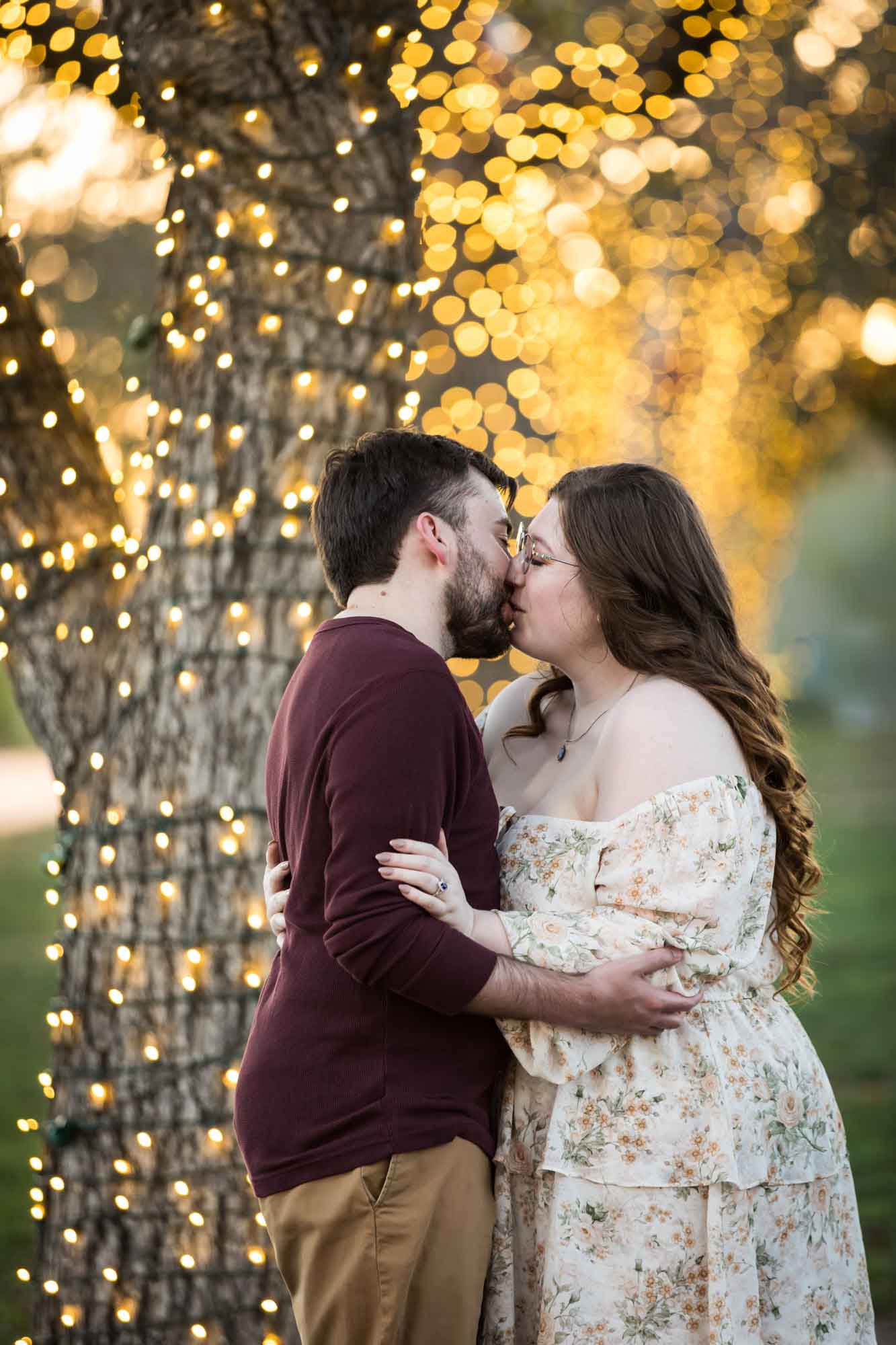 Boerne engagement photos of woman and man kissing beside trees wrapped in string lights in Main Plaza