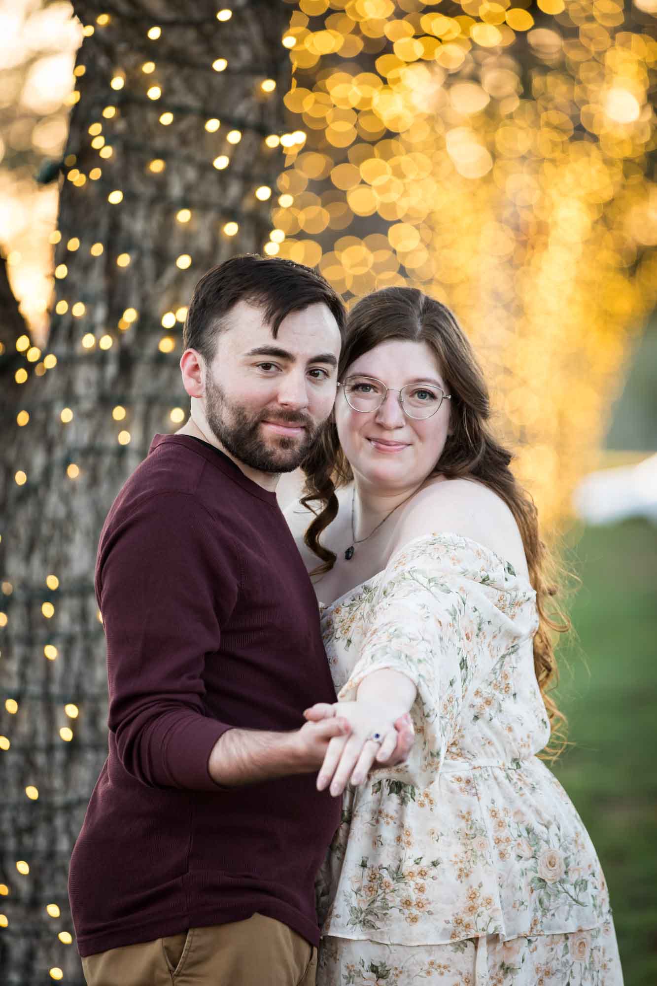 Boerne engagement photos of woman and man hugging beside trees wrapped in string lights in Main Plaza