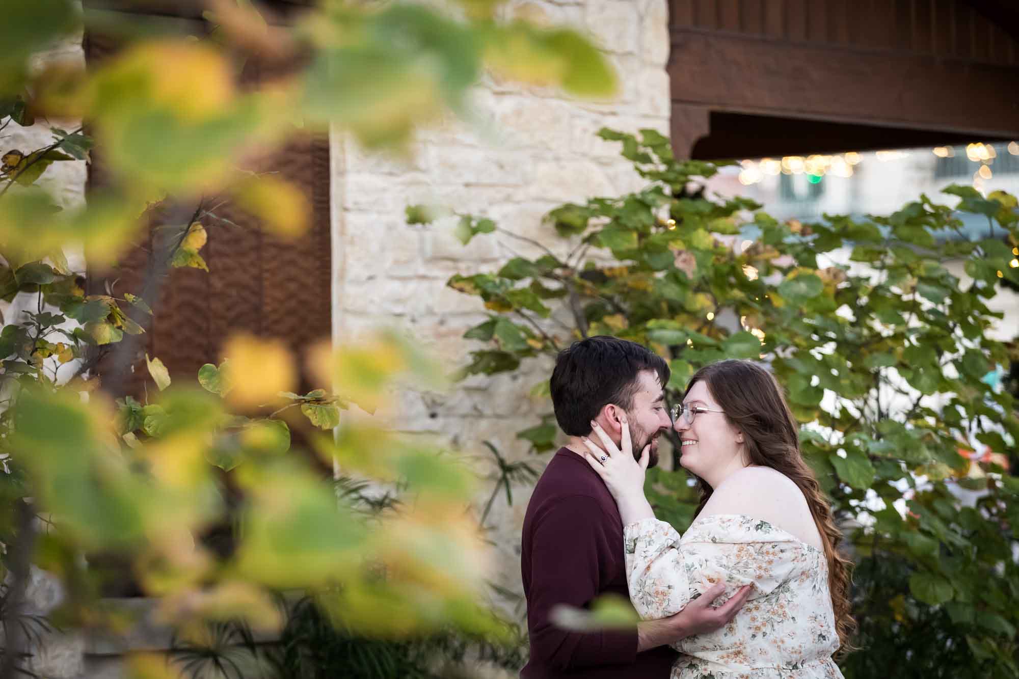 Boerne engagement photos of woman and man hugging in front of stone wall through tree branches on Main Street