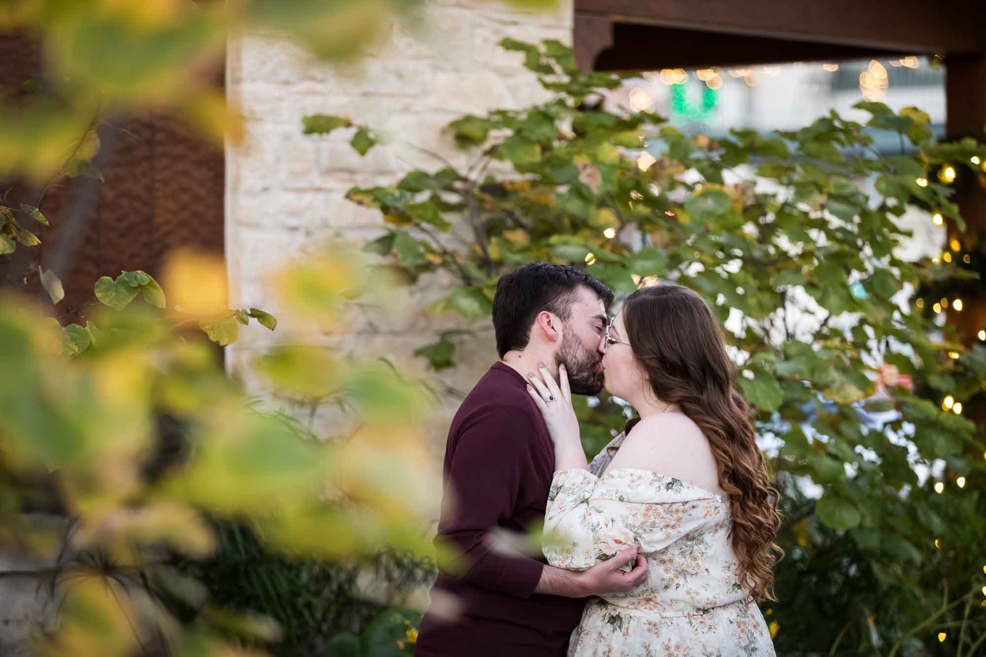 Boerne engagement photos of woman and man kissing in front of stone wall through tree branches on Main Street