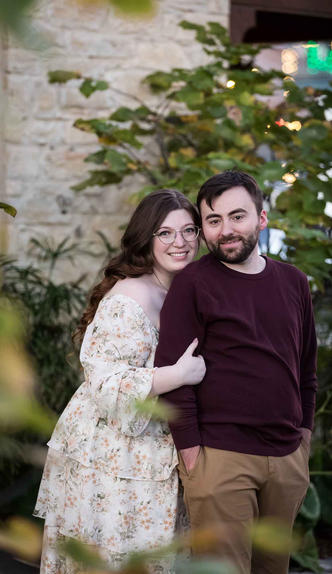 Boerne engagement photos of woman and man hugging in front of stone wall through tree branches on Main Street
