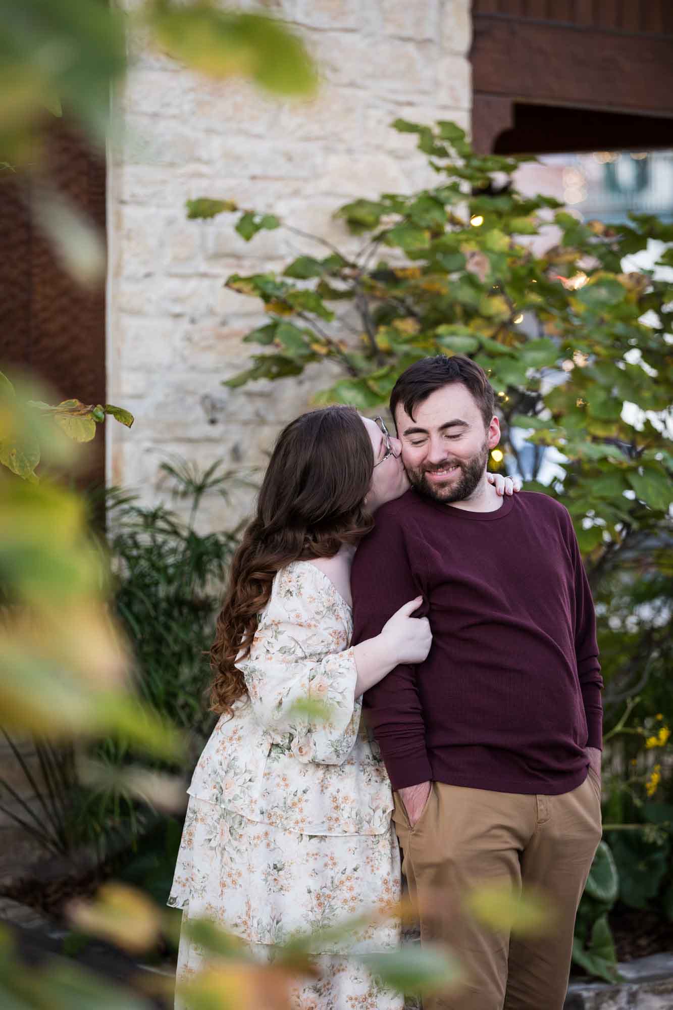 Boerne engagement photos of woman and man kissing in front of stone wall through tree branches on Main Street