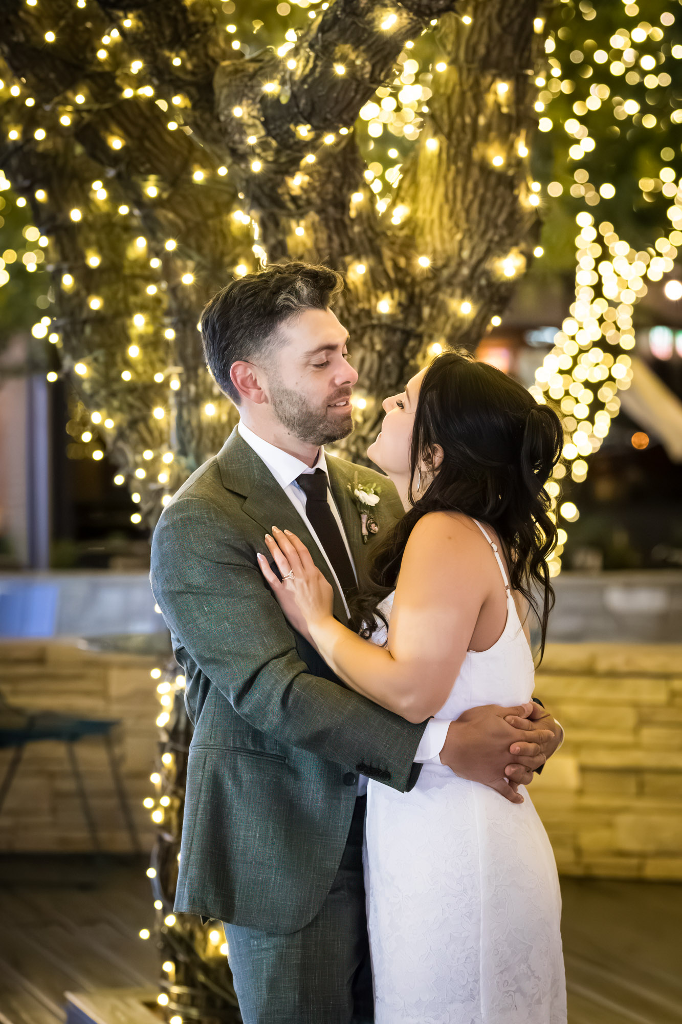 Bride and groom hugging in front of tree wrapped in string lights outside